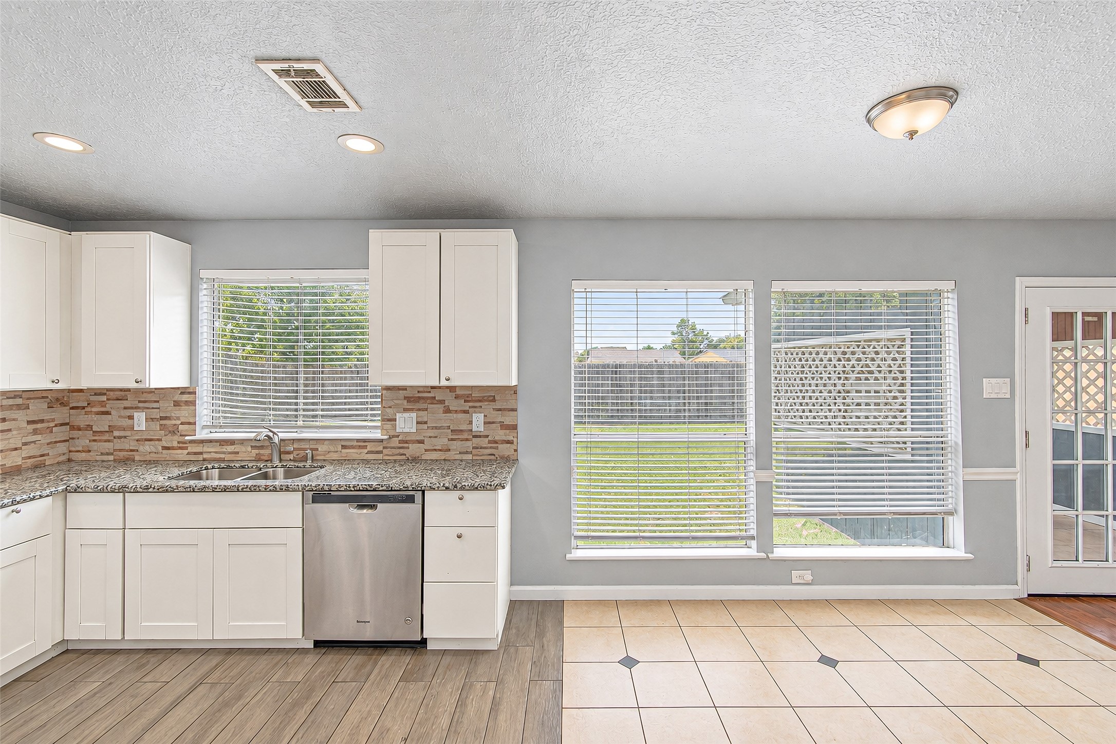 Bright kitchen and dining area featuring modern white cabinets, granite countertops, and a stylish tile backsplash, with ample natural light from large windows overlooking a spacious backyard.