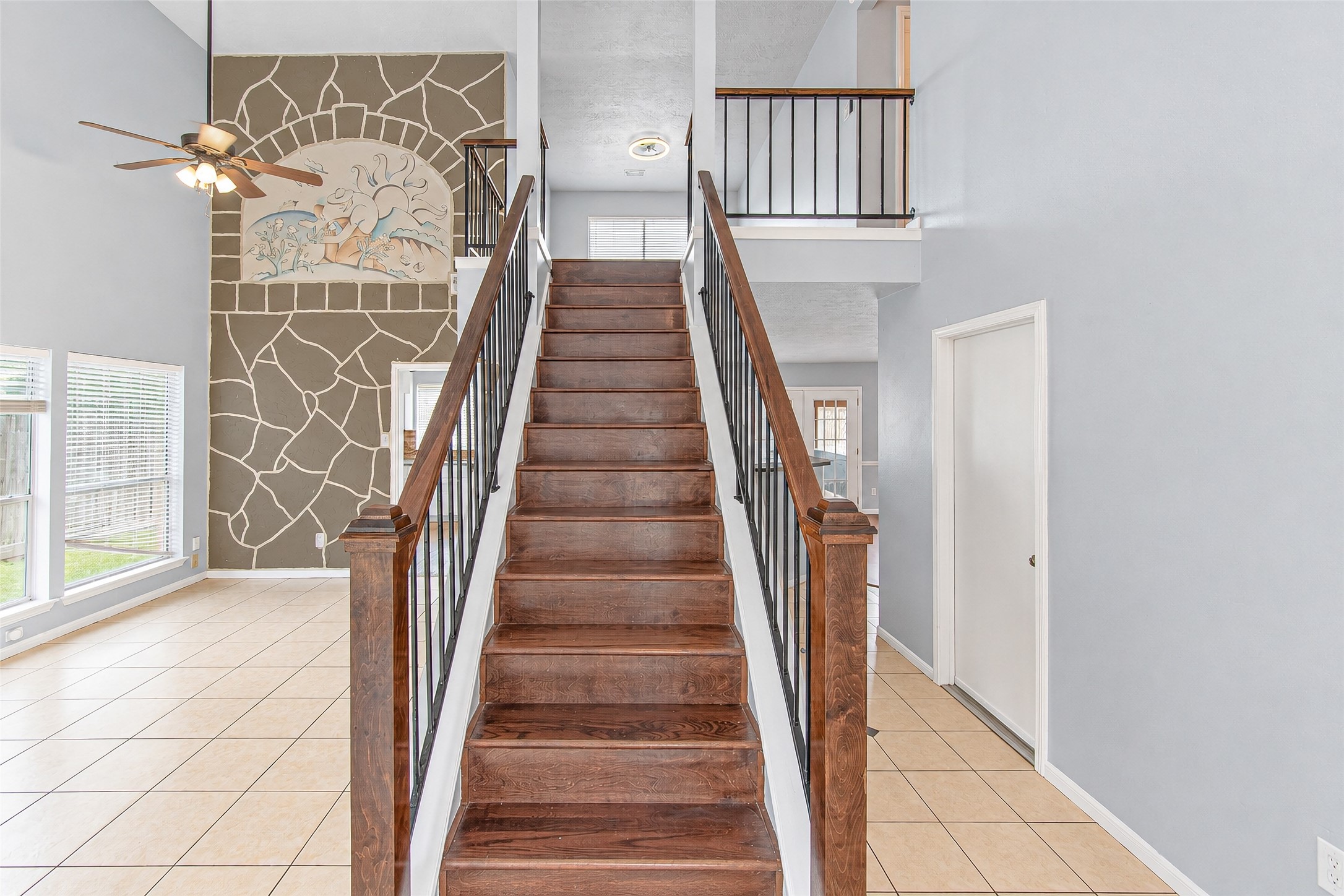 Elegant foyer featuring a stunning wooden staircase with iron railings, leading to an airy loft space. The high ceiling and large windows fill the area with natural light, and a unique stone accent wall adds character.
