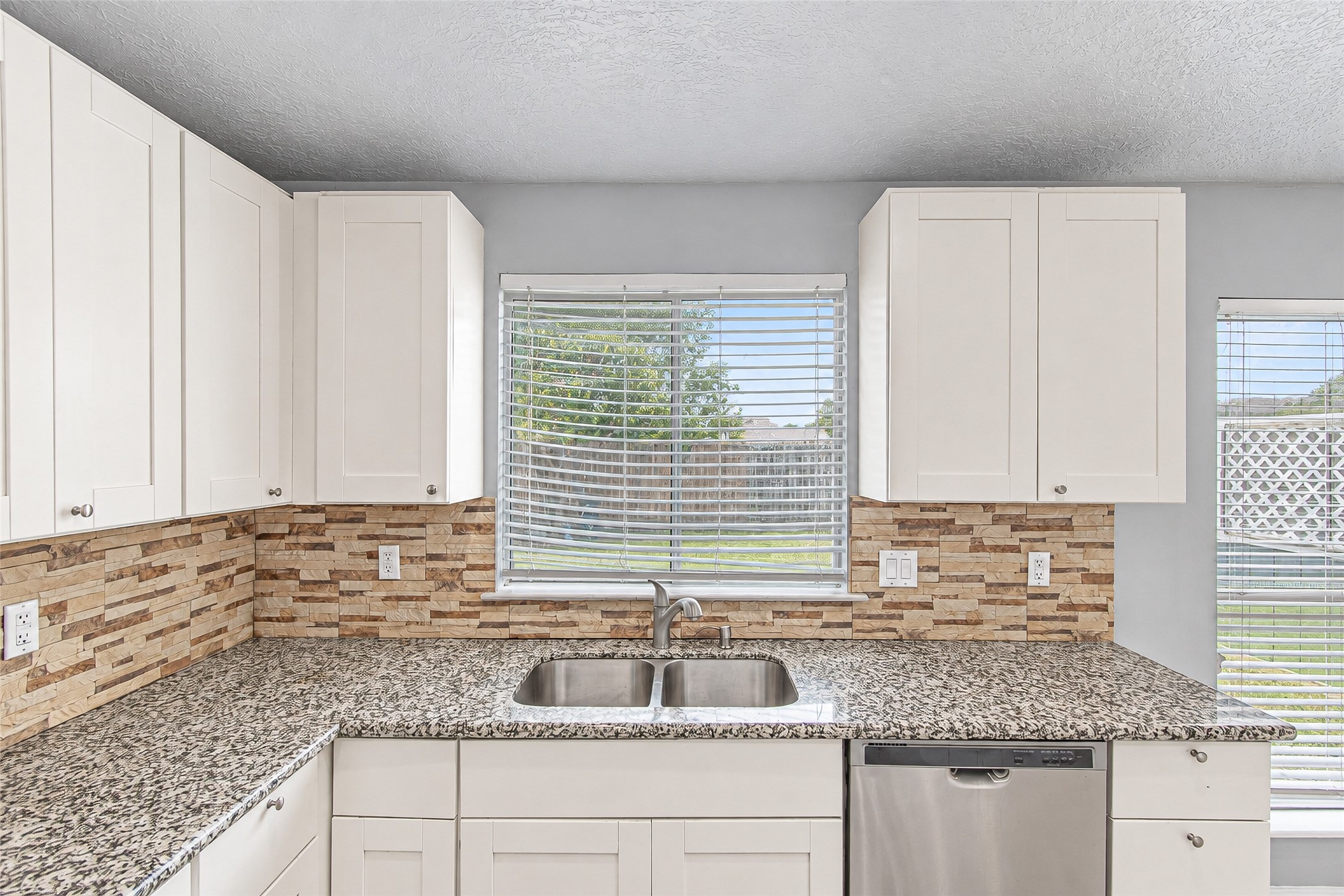 Bright kitchen featuring sleek granite countertops, modern stainless steel appliances, and ample white cabinetry, complemented by a stylish tile backsplash and a window view.