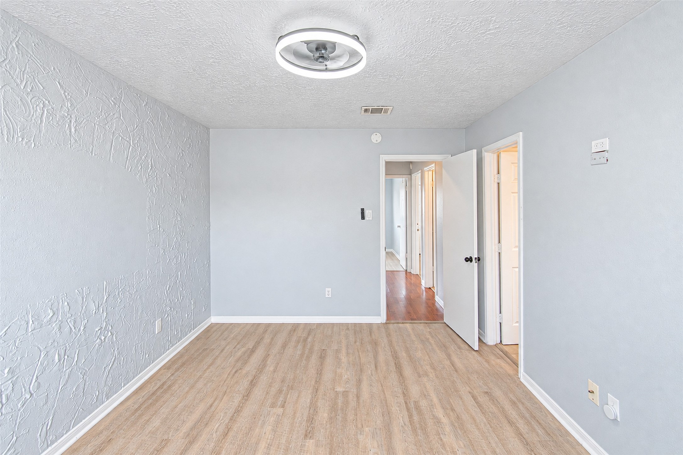 Bright room with textured walls and wood-like flooring, featuring a modern ceiling light and access to adjoining spaces through white doors.