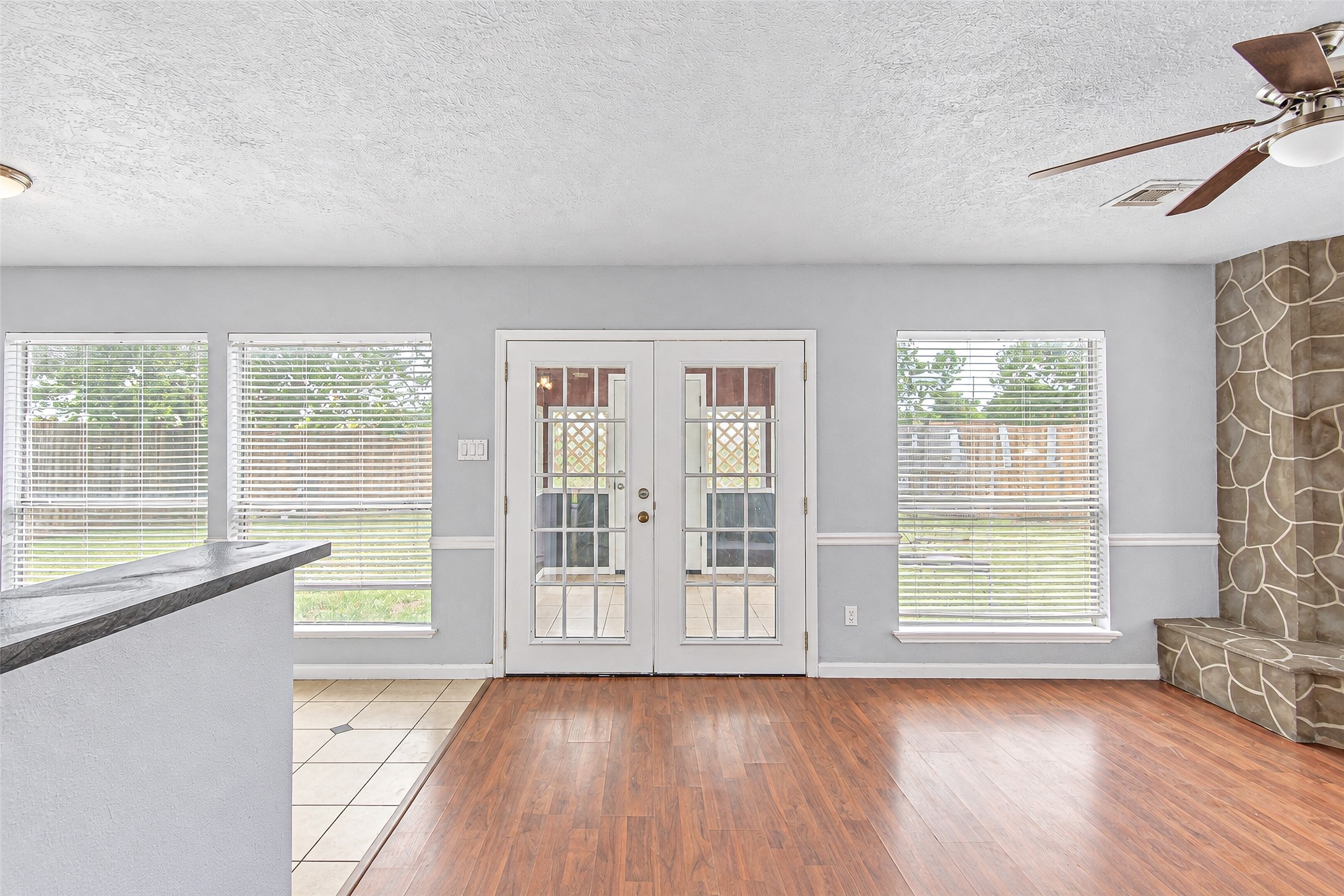 Bright living space with hardwood floors, large windows, and French doors opening to the backyard, featuring a unique stone accent wall and ceiling fan.