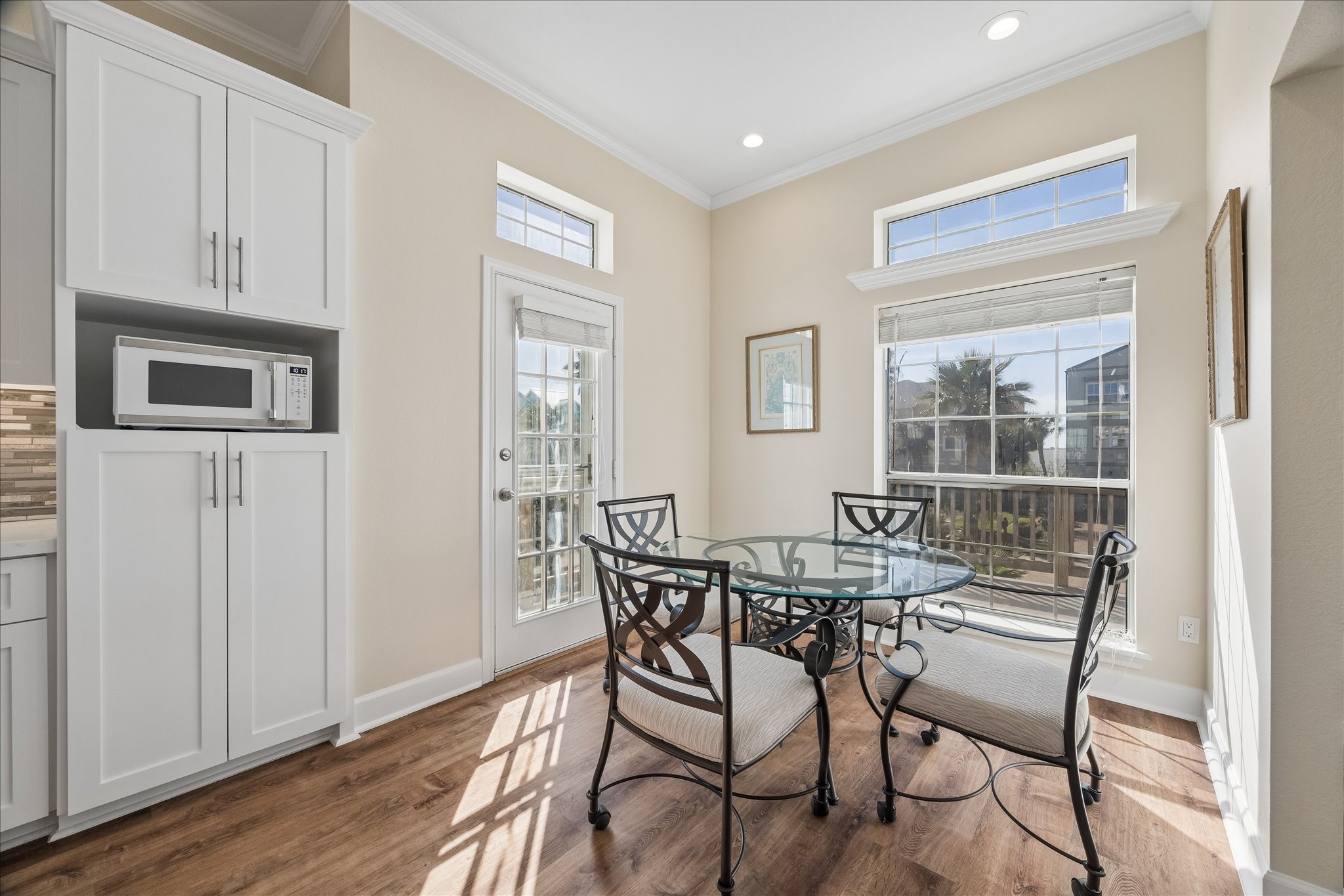 This bright dining area features large windows, a glass-top table with elegant chairs, and access to a patio through a door with glass panels. The adjacent cabinetry provides storage and includes a built-in microwave. The space is enhanced by natural light and wood flooring.