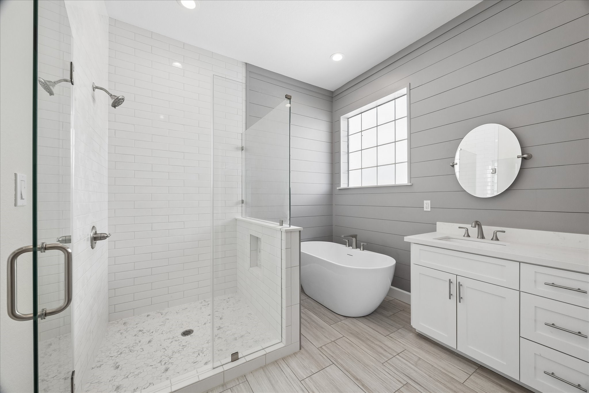 This bathroom features a modern design with a spacious glass-enclosed shower, a freestanding bathtub, and a double vanity with sleek fixtures. The room is brightened by a large window and has elegant gray shiplap walls and light wood flooring.