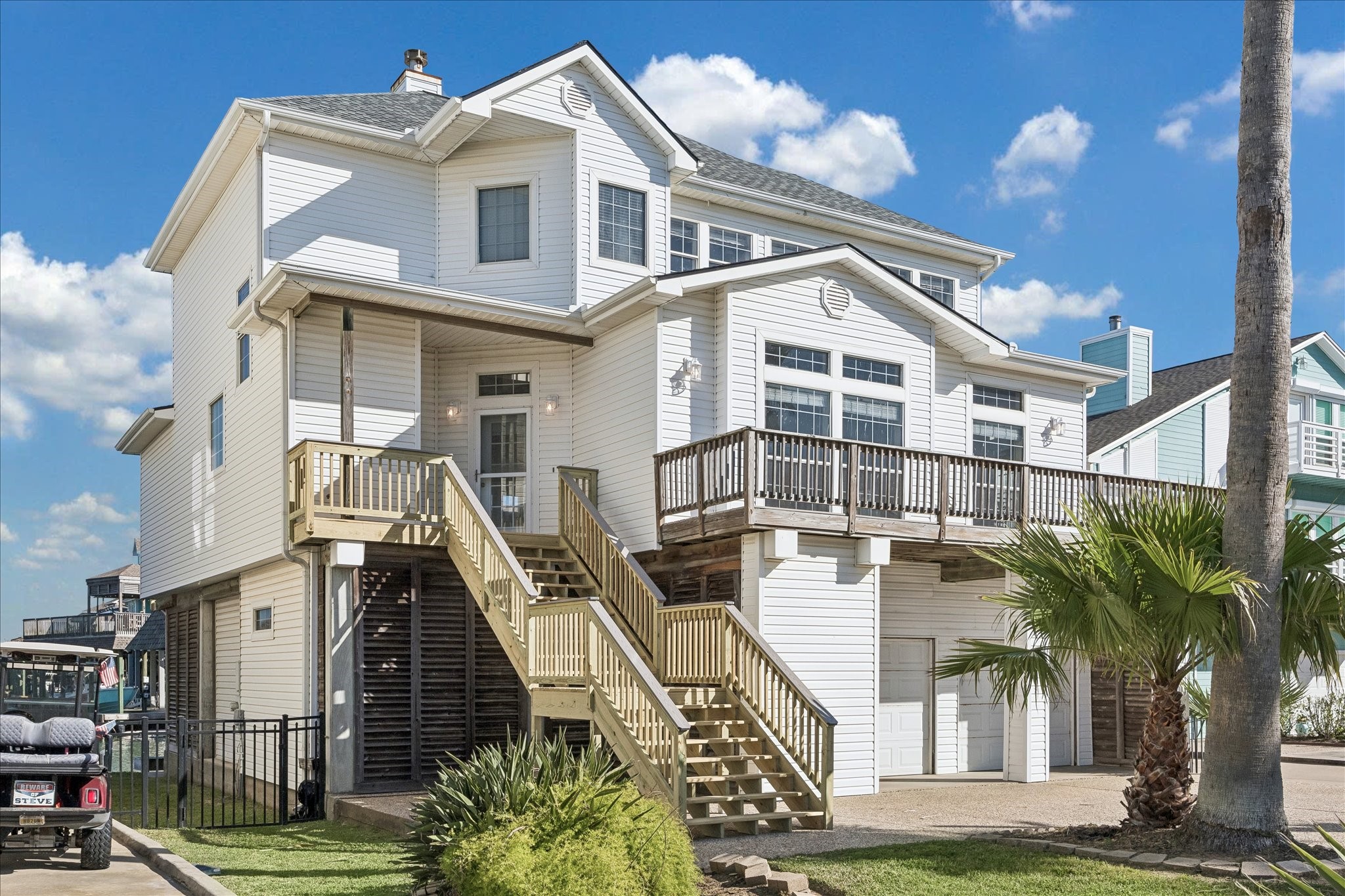 This is a spacious, elevated two-story white home with a large balcony and an exterior staircase. It features a two-car garage and is surrounded by a well-maintained landscape, including palm trees, under a clear blue sky. Ideal for coastal living.