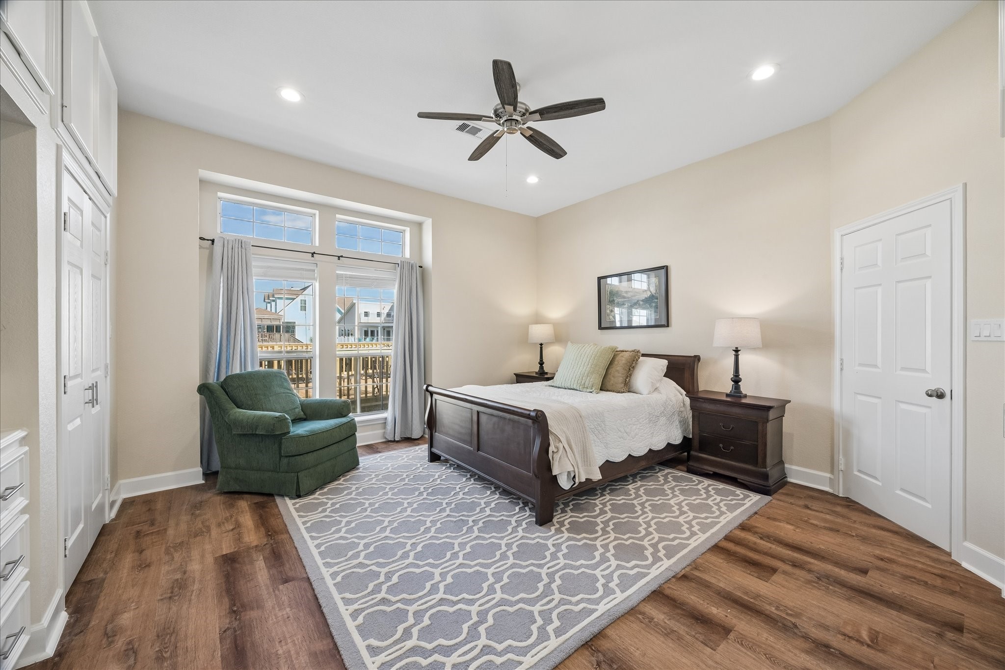 Bright and spacious bedroom with wood flooring, featuring a large bed, cozy green armchair, and elegant decor. Natural light floods in through the ample windows, complemented by a ceiling fan and recessed lighting.