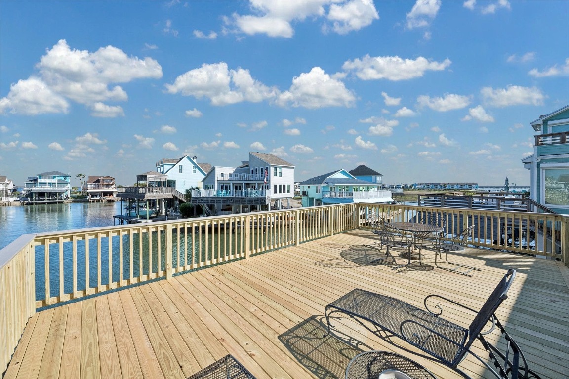 This image showcases a spacious waterfront deck with wooden railings and outdoor seating, offering stunning views of nearby waterfront homes and a serene body of water under a clear, blue sky. Perfect for relaxation and entertaining.