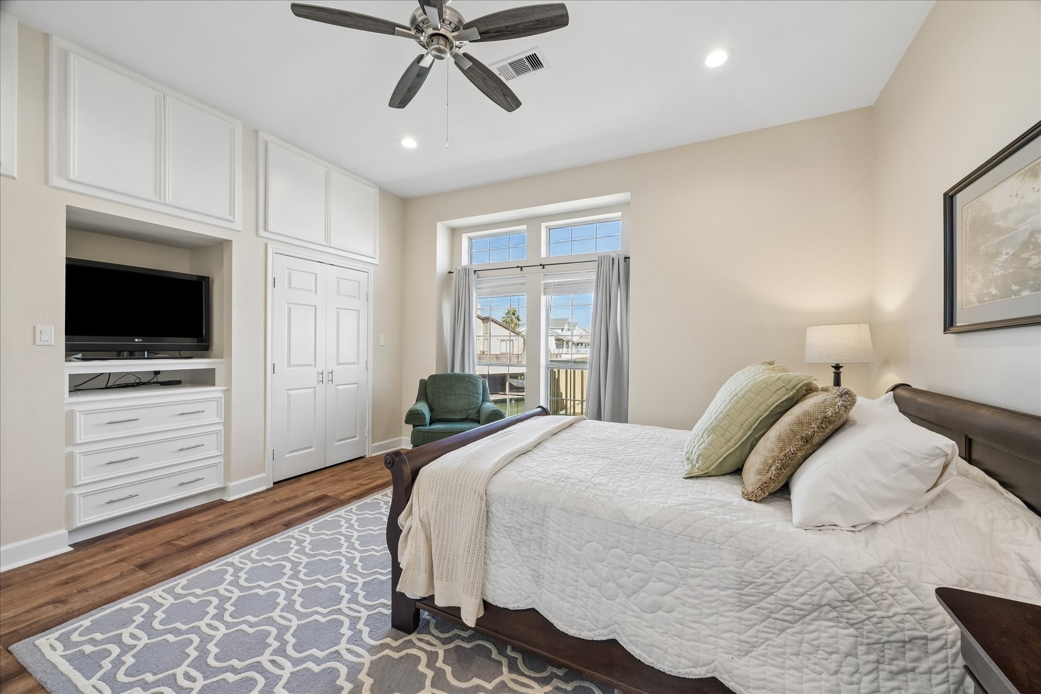 This cozy bedroom features a large window for natural light, a ceiling fan, built-in storage, a TV, and hardwood floors. It includes a comfortable bed with stylish bedding and a tasteful area rug, creating a welcoming atmosphere. This cozy bedroom features a large window for natural light, a ceiling fan, built-in storage, a TV, and hardwood floors. It includes a comfortable bed with stylish bedding and a tasteful area rug, creating a welcoming atmosphere.