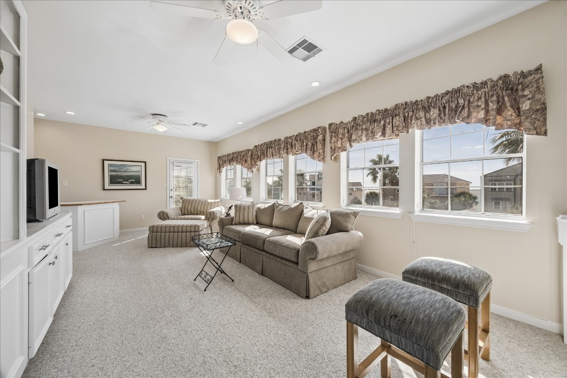 Bright and spacious living room featuring large windows with scenic views, neutral carpet, and cozy seating. Includes built-in shelving and a ceiling fan for comfort and convenience.