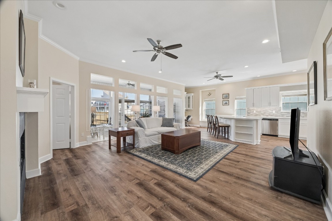 This spacious open-concept living area features elegant wood flooring and abundant natural light. It includes a cozy seating area with a fireplace, a modern kitchen with white cabinetry and a large island, and a dining space, perfect for entertaining. Large windows and ceiling fans enhance the bright and airy atmosphere.