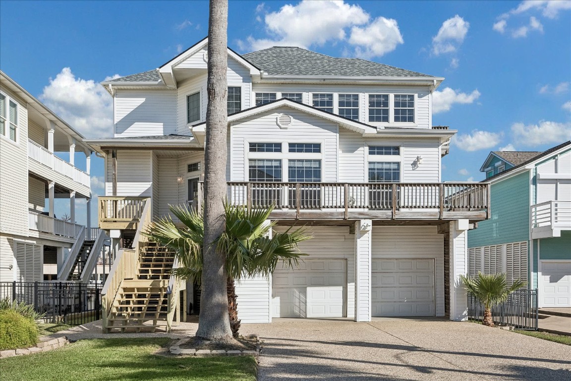 This is a charming, multi-story coastal home with a white exterior, featuring expansive windows and a large front deck. It has a double garage and is elevated with a stairway entrance, complemented by palm trees and a well-maintained front yard. This is a charming, multi-story coastal home with a white exterior, featuring expansive windows and a large front deck. It has a double garage and is elevated with a stairway entrance, complemented by palm trees and a well-maintained front yard.