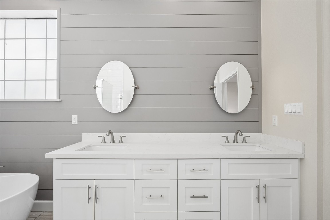Modern bathroom features a double vanity with white cabinetry and sleek faucets, set against a gray shiplap wall. Two round mirrors and a window provide ample light, creating a bright, inviting space.