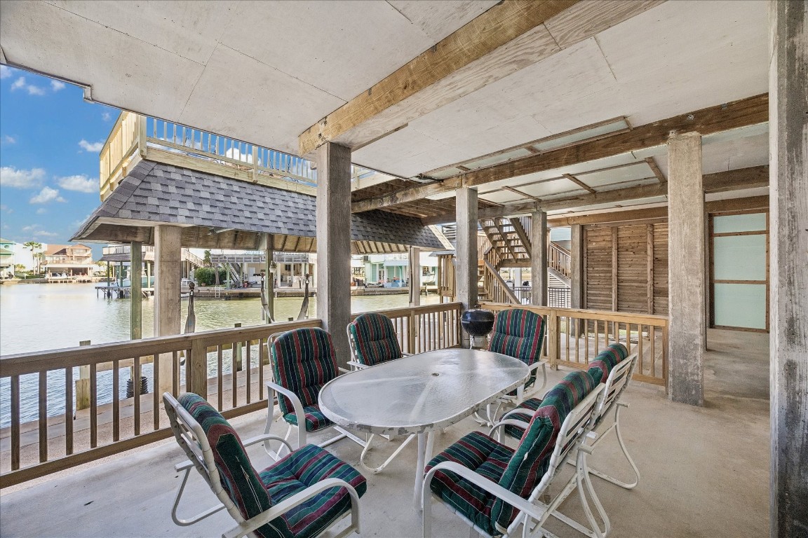 This photo showcases a covered patio with a waterfront view, perfect for outdoor dining and relaxation. It features a table with seating, surrounded by a wooden railing, and offers scenic views of the canal and nearby homes.