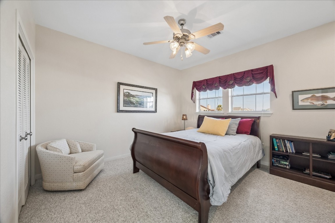 This cozy bedroom features a plush carpet, a classic wooden bed, and a ceiling fan. It has a window with a decorative valance, a comfortable armchair, and a small bookshelf. The neutral walls are accented with framed art, creating a welcoming atmosphere.