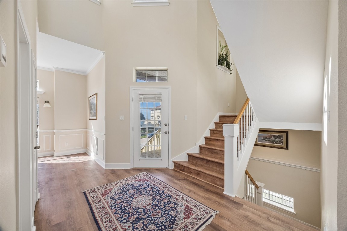 This welcoming entryway features high ceilings, abundant natural light, and elegant wood flooring. A stylish staircase leads to the upper level, while a glass door and tasteful wainscoting add charm.