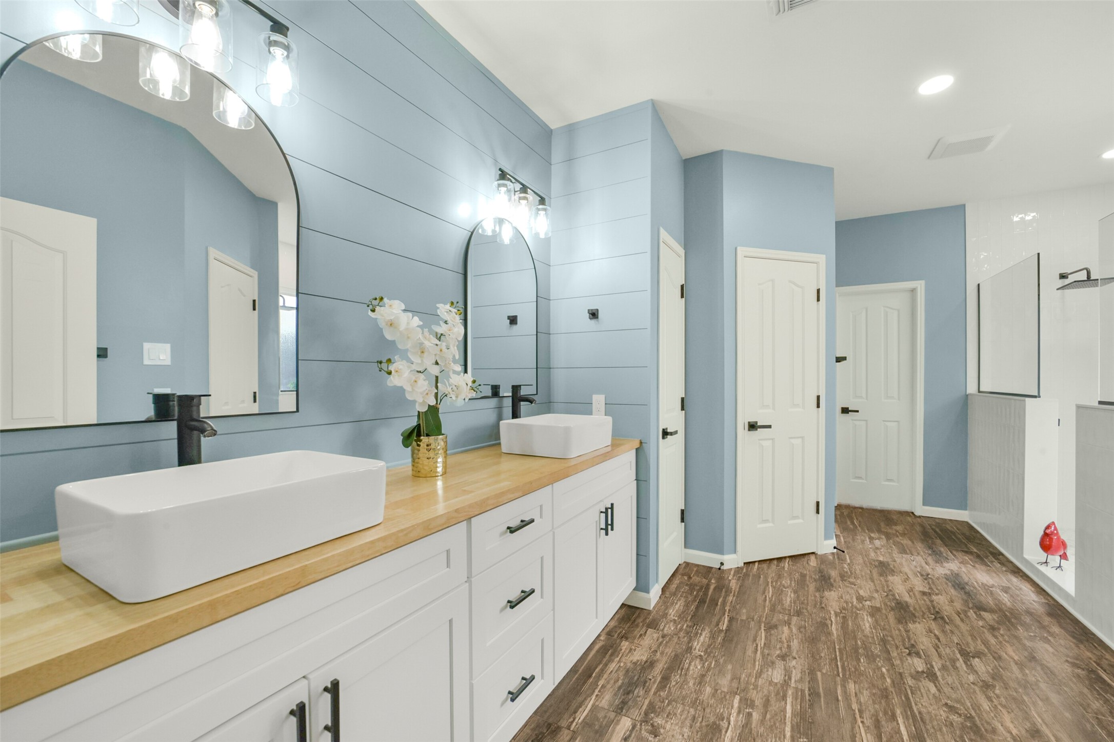 Elegant bathroom featuring dual vessel sinks on a sleek wooden countertop, complemented by modern black fixtures and stylish lighting. The blue shiplap walls and wood-look flooring create a serene, spa-like ambiance.