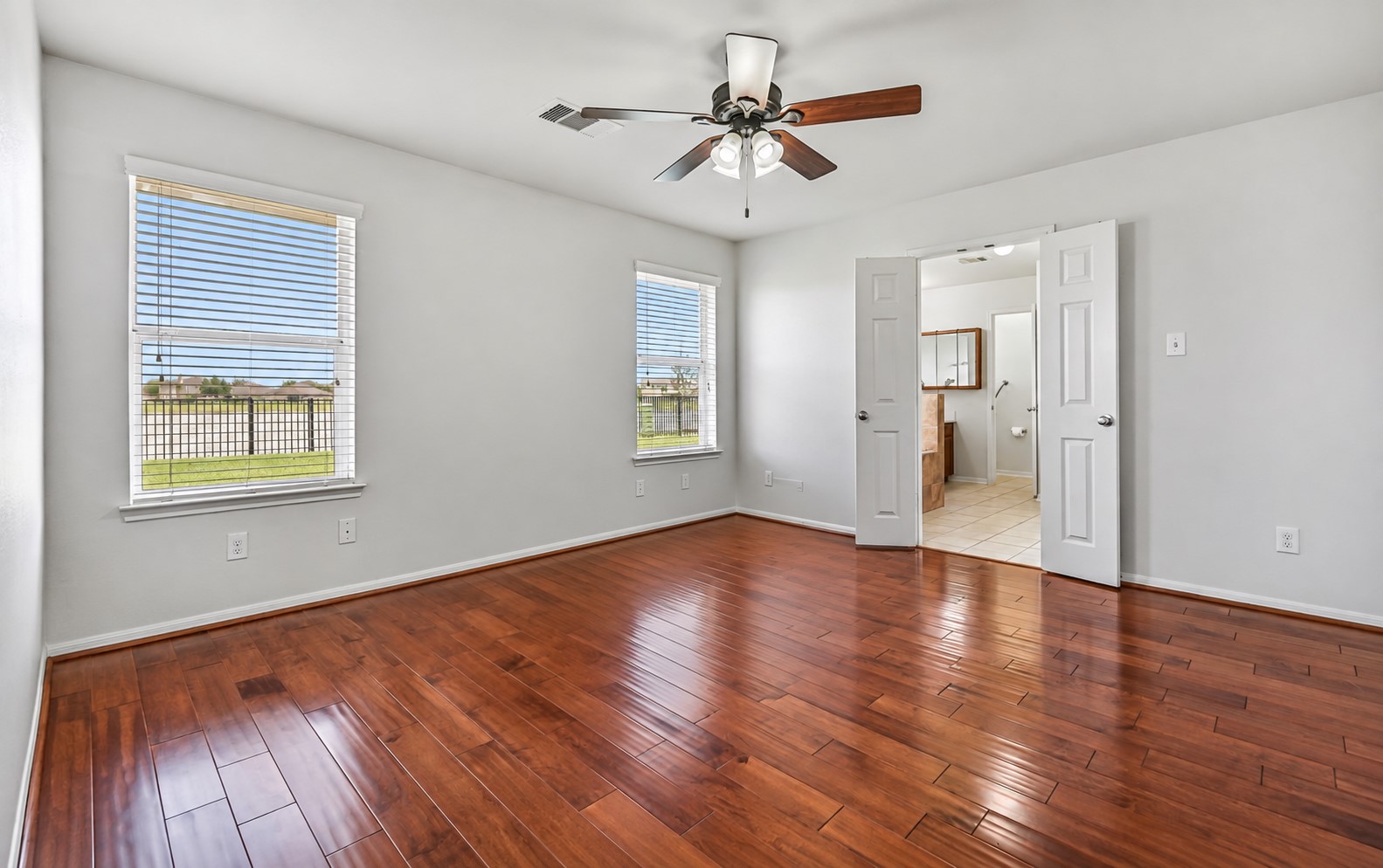 The secluded primary suite offers a peaceful retreat with rich wood flooring, fresh neutral paint, ceiling fan & two large windows framing views of the backyard and pond, while double doors lead directly into the spacious en-suite bath.