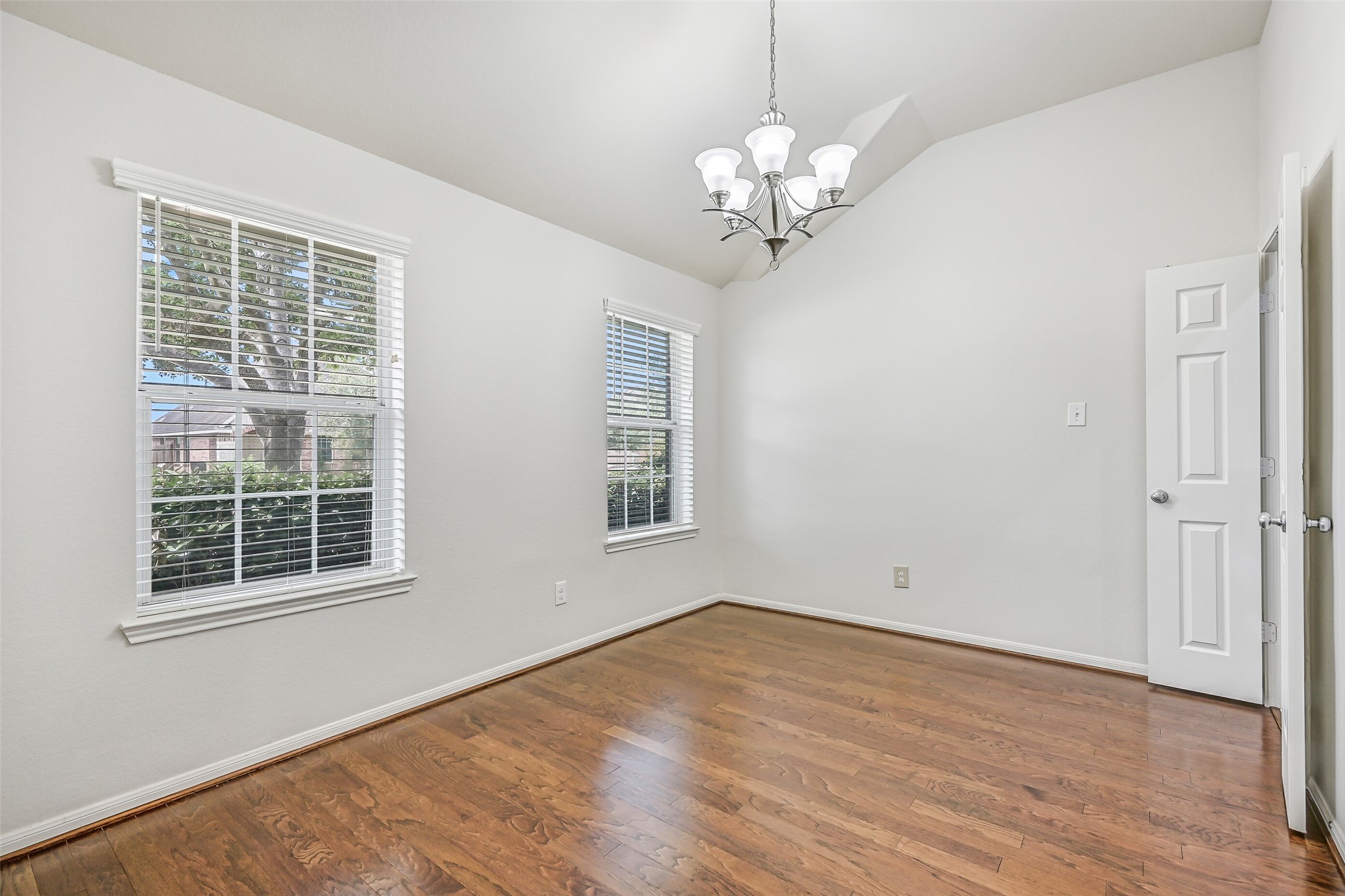 A second view of the light-filled dining room highlights dual windows with 2" blinds, beautiful wood flooring & the soaring vaulted ceiling, offering generous space for both everyday meals and special gatherings.