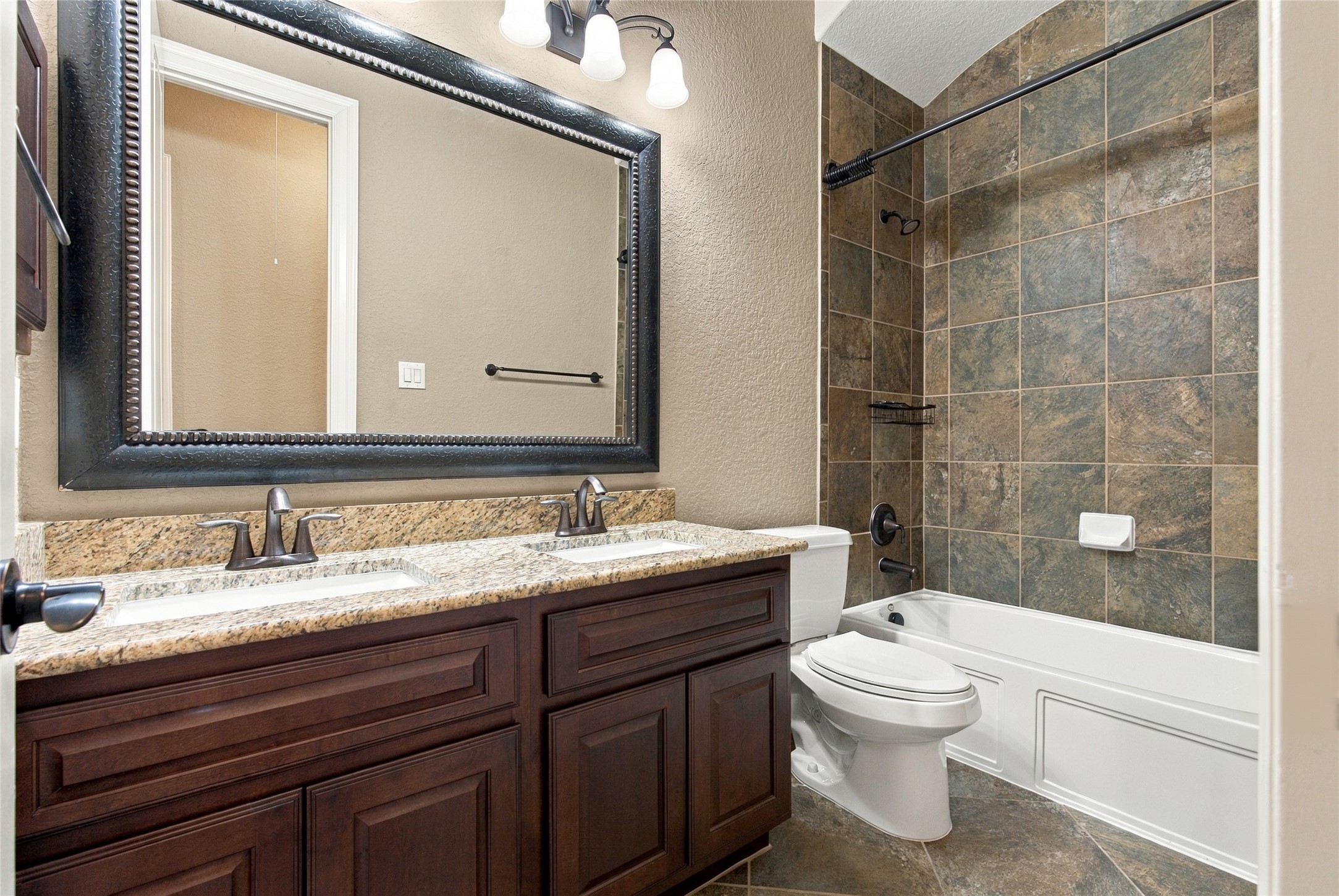 Full downstairs secondary bathroom featuring a granite countertop, double sinks, framed mirror, and tub/shower combination with decorative tile surround.