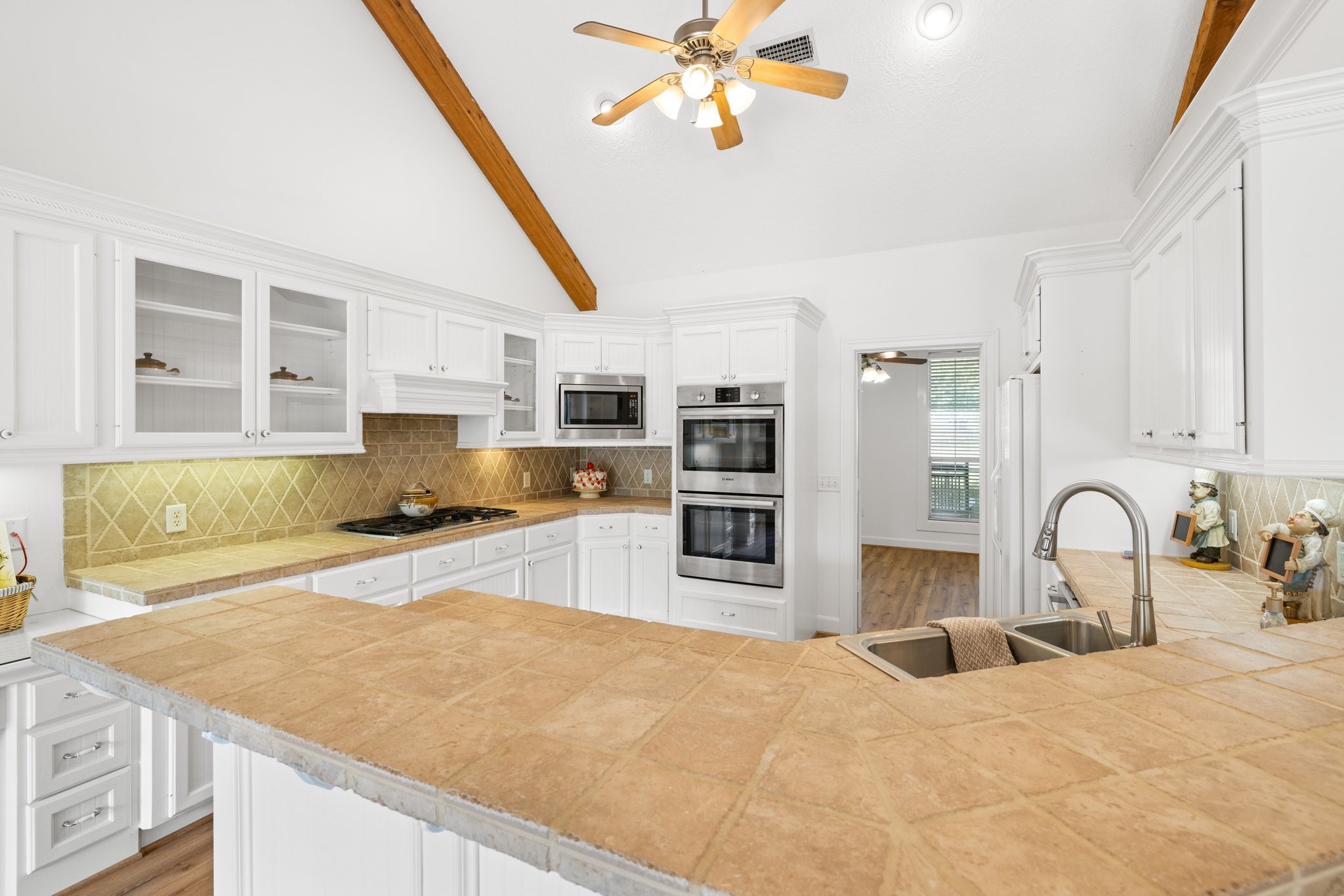 This kitchen features high vaulted ceilings with exposed wooden beams, white cabinetry, and a central island with a breakfast bar. It includes stainless steel appliances, a double oven, and a modern sink, all complemented by warm tile countertops and backsplash.