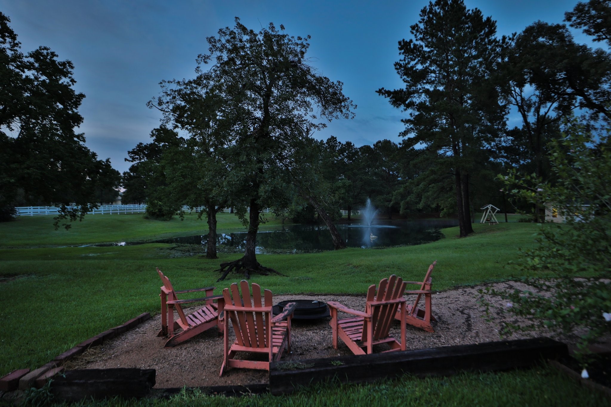 This photo showcases a serene property at dusk, featuring a spacious yard with mature trees, a tranquil pond with a fountain, and room for your cozy seating around a fire pit. Perfect place to unwind with your guests!