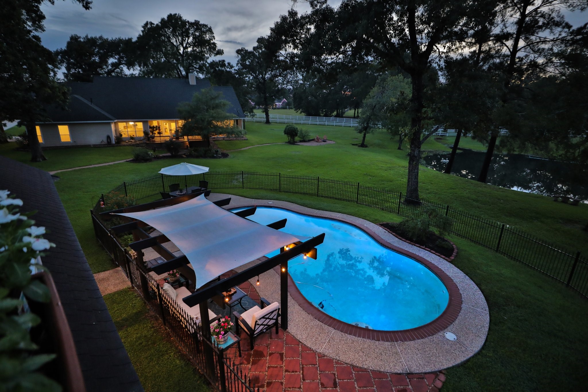 This aerial view shows a spacious backyard with a serene pool, shaded pergola area, and lush greenery, ideal for relaxation and entertaining. The house is warmly lit and offers a picturesque view of the pond and surrounding trees, creating a tranquil and inviting atmosphere.