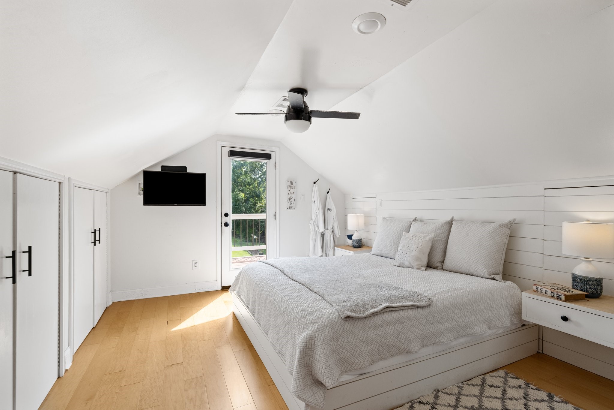 This cozy bedroom features a sloped ceiling, beautiful floors and accent wall. It has built-in storage,  and a glass door leading to an outdoor balcony, creating a bright and inviting space.