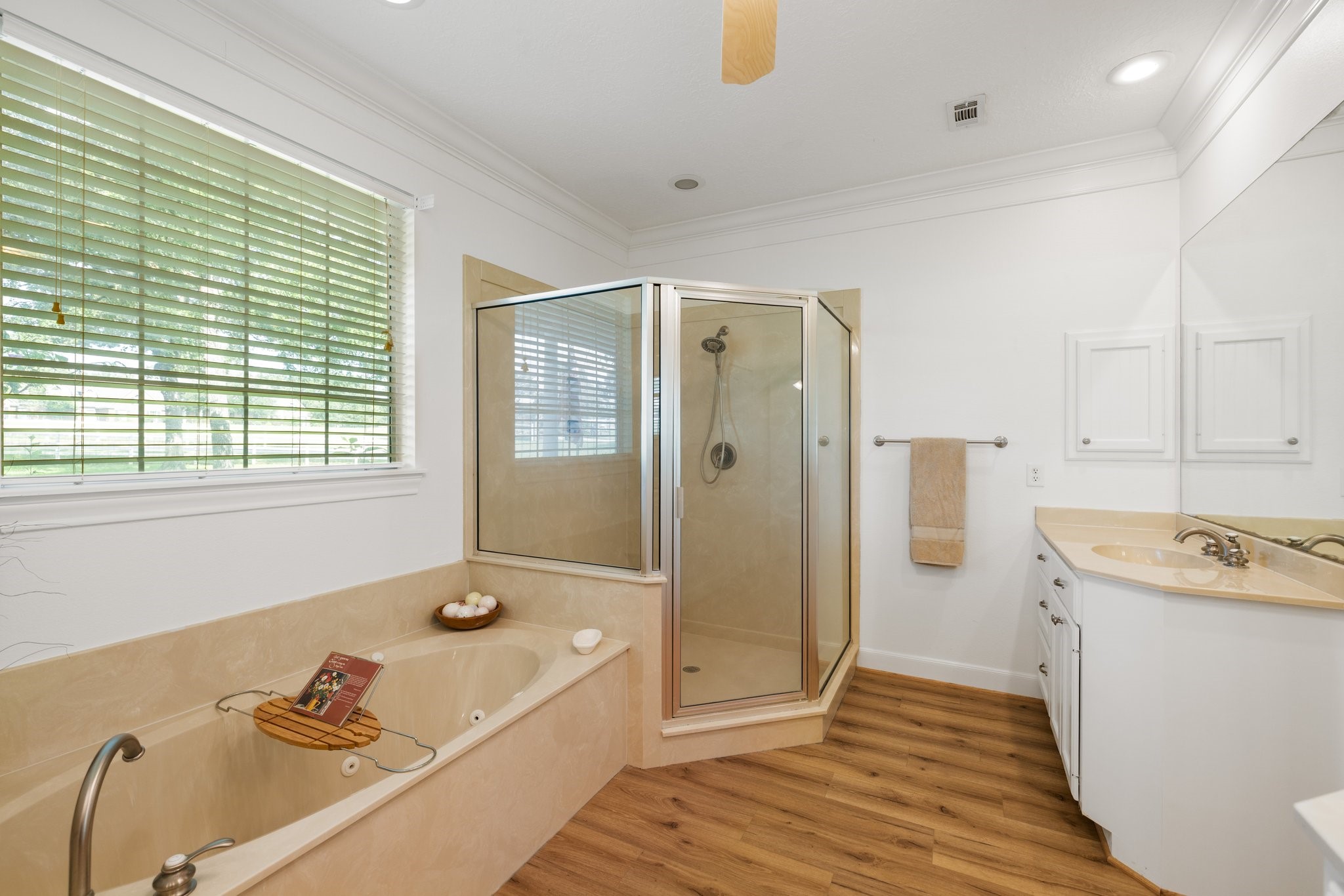 This bathroom features a spacious layout with a large jetted soaking tub, a separate glass-enclosed shower, and a dual-sink vanity. Natural light streams in through a window with blinds, highlighting the neutral color palette for a modern, inviting atmosphere.