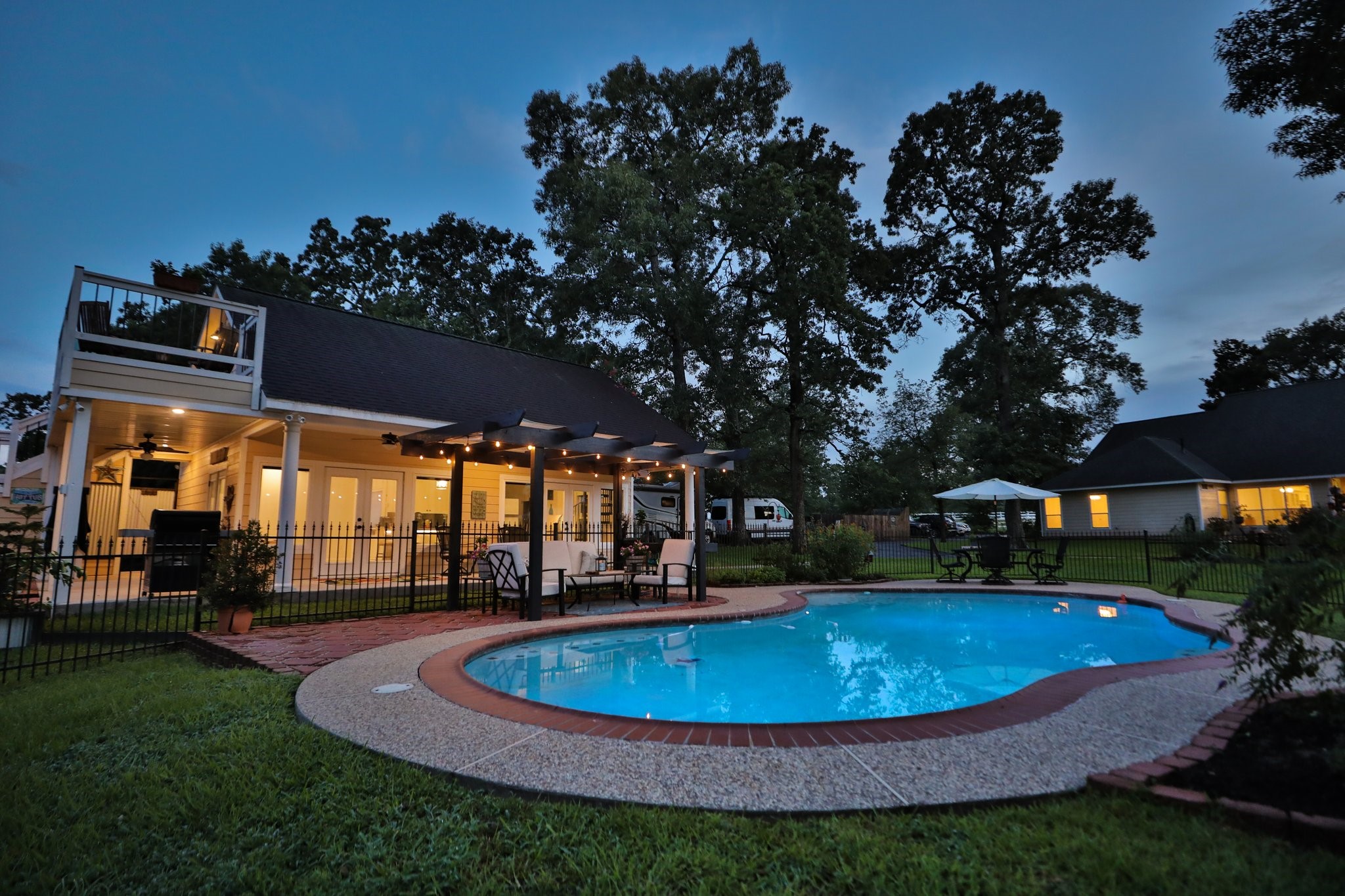 This photo showcases a charming backyard featuring a well-lit pool with surrounding patio space and guest house. The adjacent deck and seating area under a pergola offer a perfect spot for relaxation or entertaining. Lush greenery and mature trees provide a peaceful, private atmosphere, complemented by the warm glow from the home's exterior lighting.