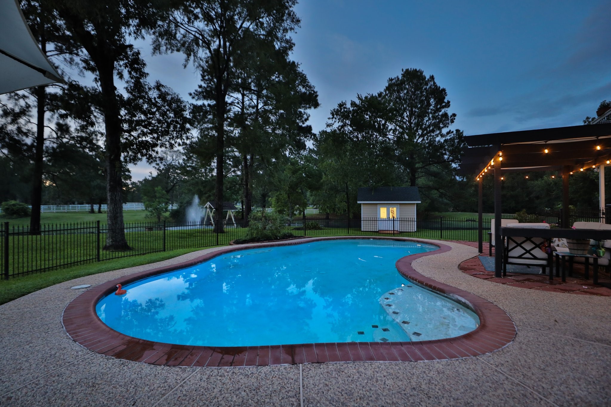 Sunset view of this inviting backyard features a well-lit swimming pool surrounded by lush greenery and a fenced-in area for privacy. A cozy pergola with seating area offers a perfect spot for relaxing or entertaining.