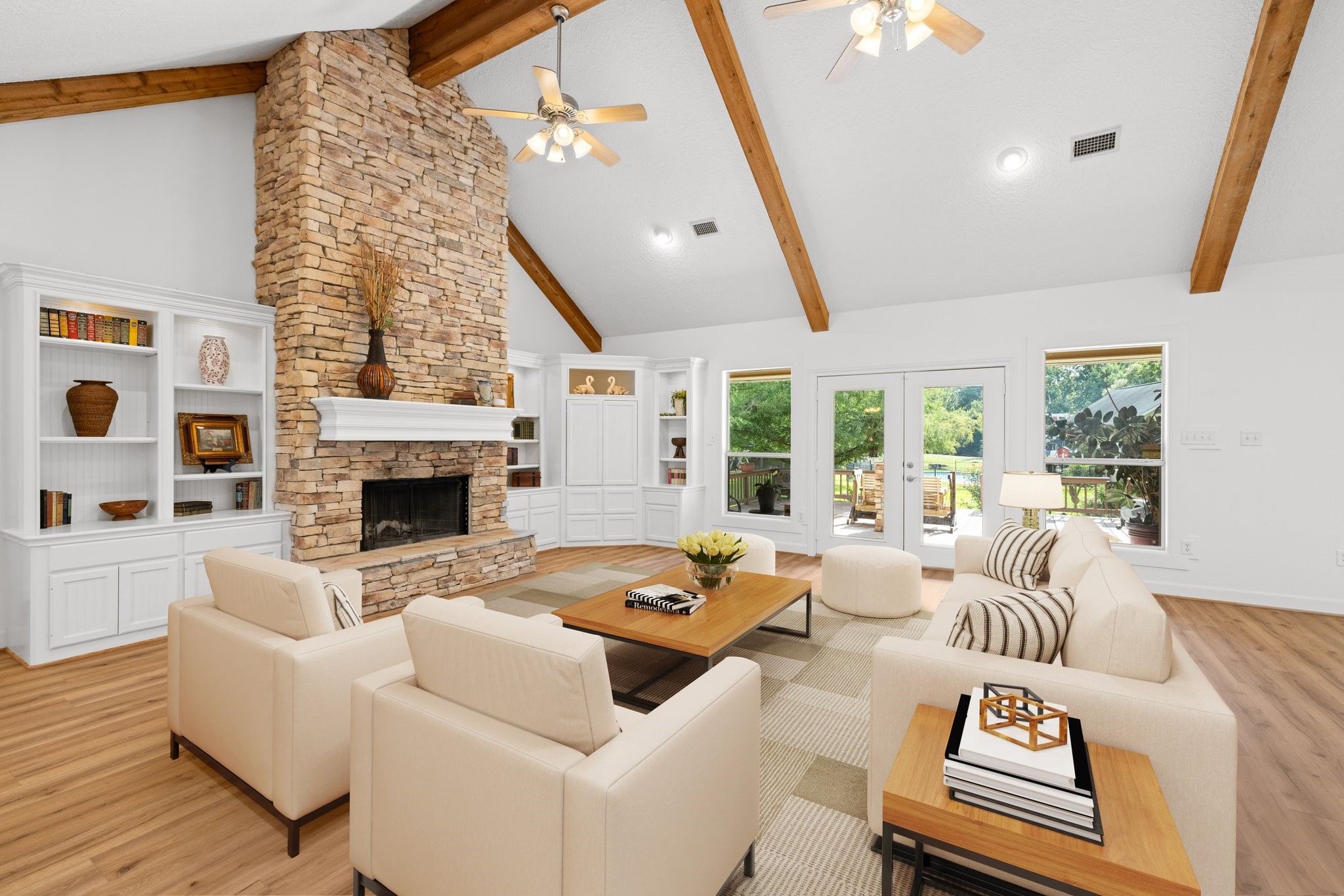 This spacious living room features a high vaulted ceiling with exposed wooden beams, a striking stone fireplace, and built-in shelving. Large windows and French doors offer plenty of natural light and views of the outdoor greenery.