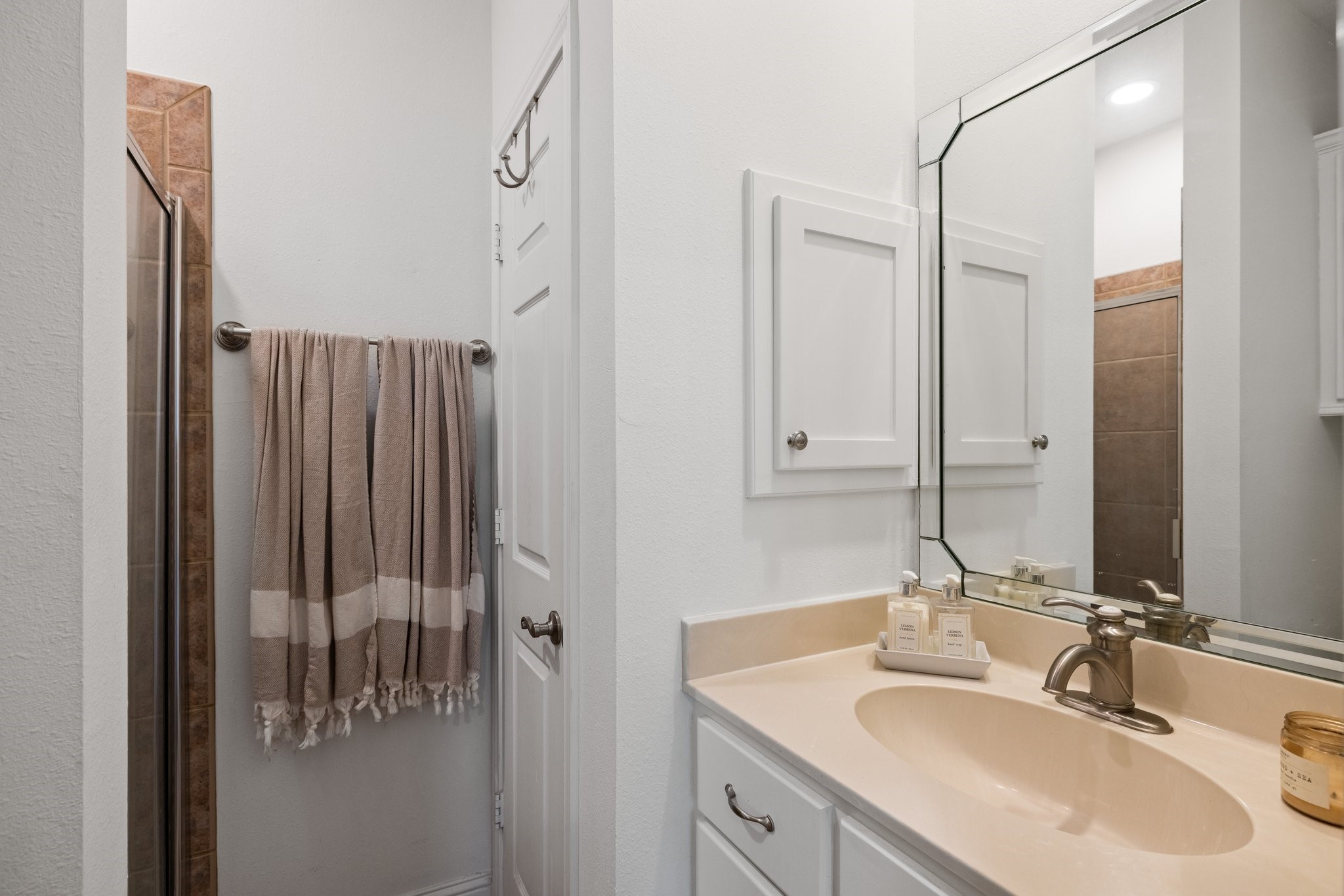 This bathroom features a modern design with a tiled shower, cream countertop, and a stylish mirror. It includes white cabinetry and a towel rack, providing a clean and functional space.