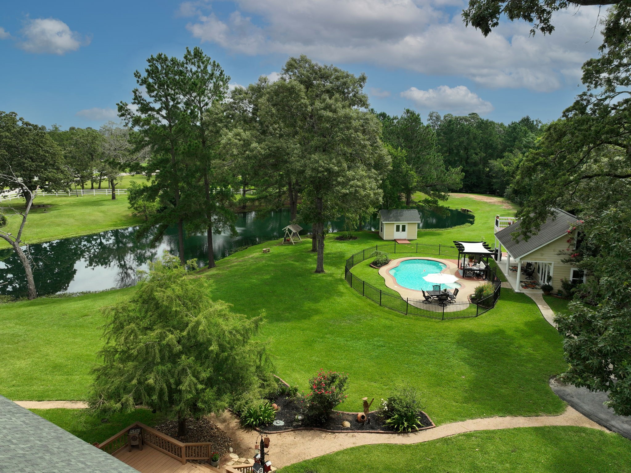 This aerial view shows lush greenery, a serene pond, and a fenced-in pool area. The charming guest house with a pergola offers a perfect spot for relaxation, surrounded by mature trees and manicured landscaping. Ideal for those seeking a peaceful retreat.