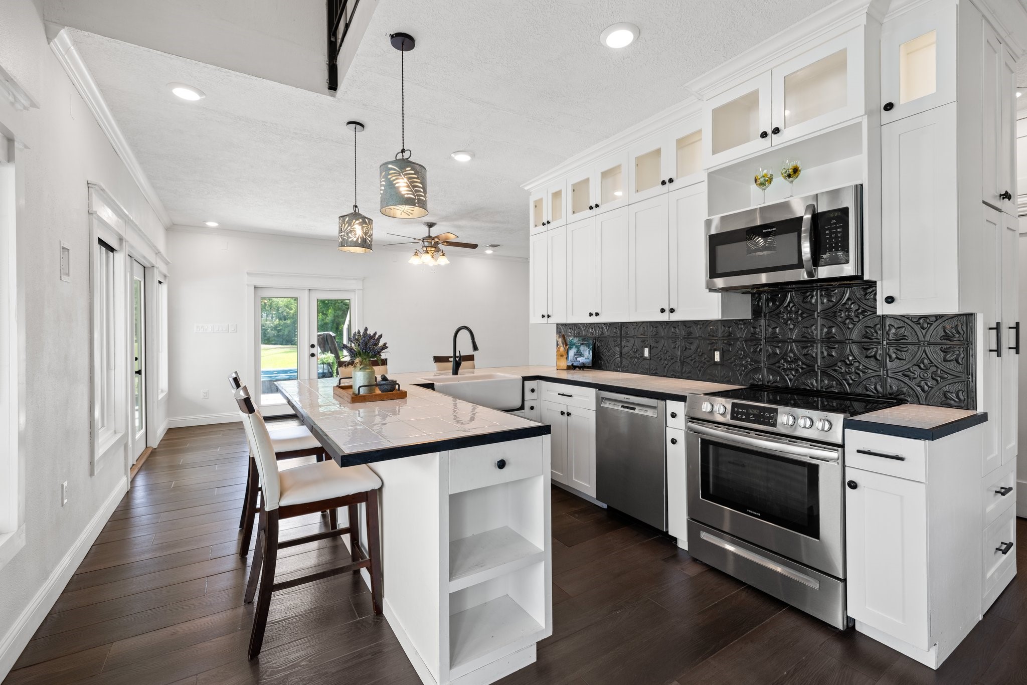 This modern kitchen fin the guest house features sleek white cabinetry with glass accents, stainless steel appliances, and a stylish black tile backsplash. The open layout includes a cozy breakfast bar, contemporary pendant lighting, and offers a spacious view to the outdoors through glass doors, creating a bright and inviting space.