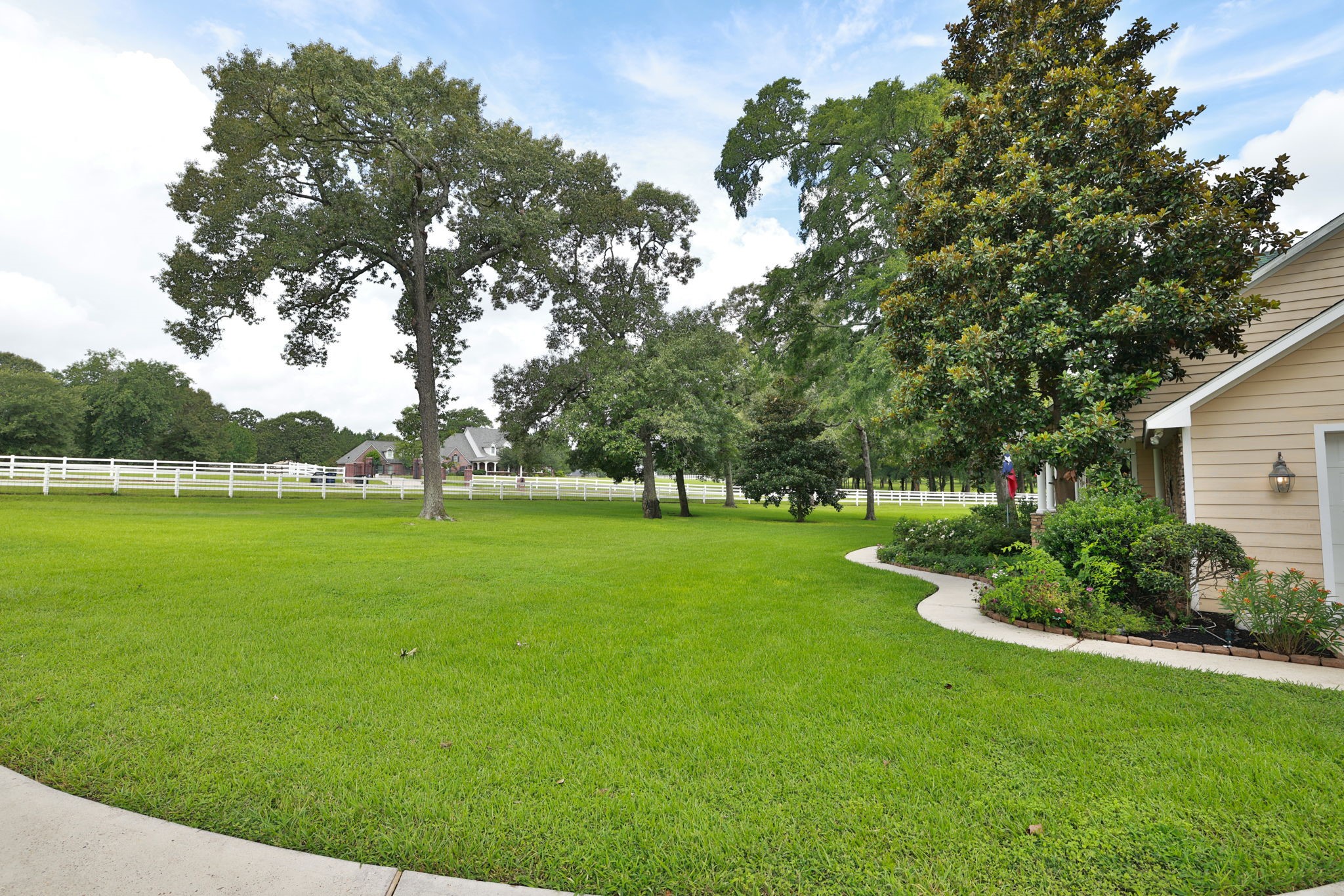 This photo showcases a spacious, well-maintained yard with lush green grass and mature trees, bordered by a white heavy gage dog/predator fence. The side of a house is visible, neatly landscaped and a curved walkway, suggesting a peaceful and inviting property. This photo showcases a spacious, well-maintained yard with lush green grass and mature trees, bordered by a white heavy gage dog/predator fence. The side of a house is visible, neatly landscaped and a curved walkway, suggesting a peaceful and inviting property.
