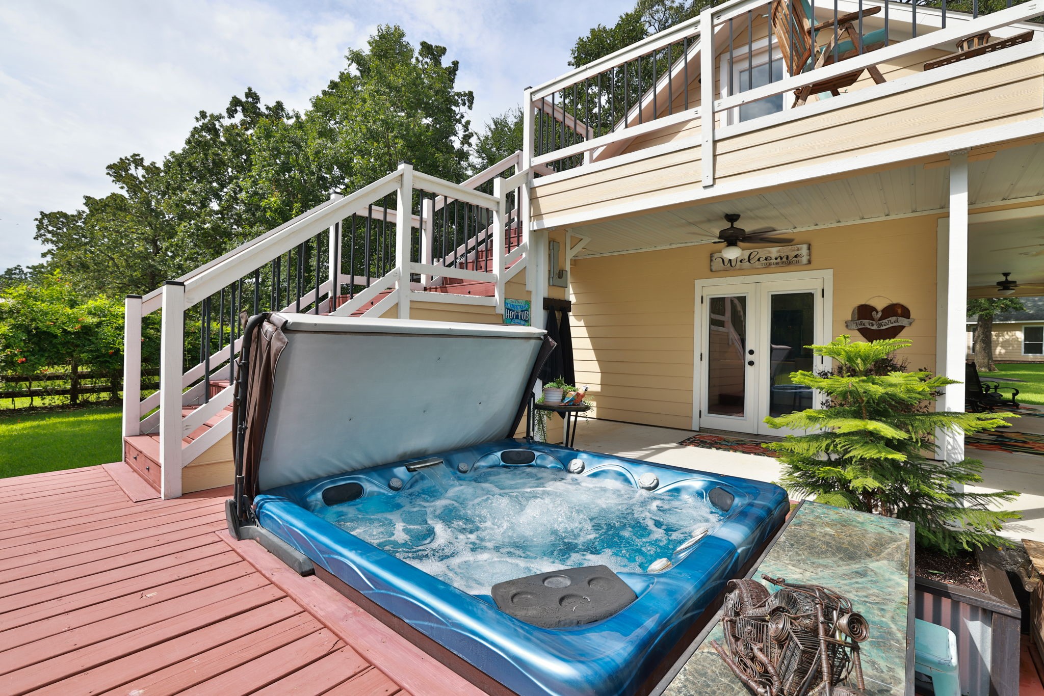 This inviting outdoor space features a bubbling hot tub on a spacious deck, leading up to a cozy balcony. The area is complemented by a charming covered patio with ceiling fans, perfect for relaxation and entertaining. Lush greenery surrounds the setting, adding to the serene atmosphere.