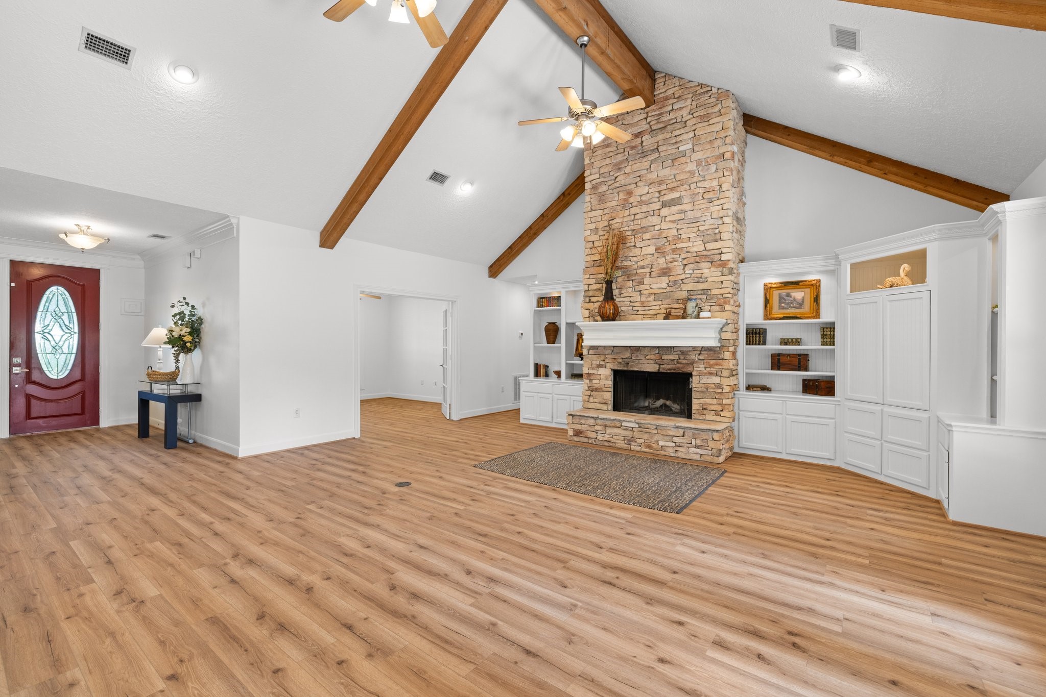 This spacious living room features a high vaulted ceiling with exposed wood beams, a beautiful floor to ceiling stone fireplace, and built-in shelving. The open layout and natural light create a warm, inviting atmosphere, complemented by vinyl plank flooring and a stylish entry door.