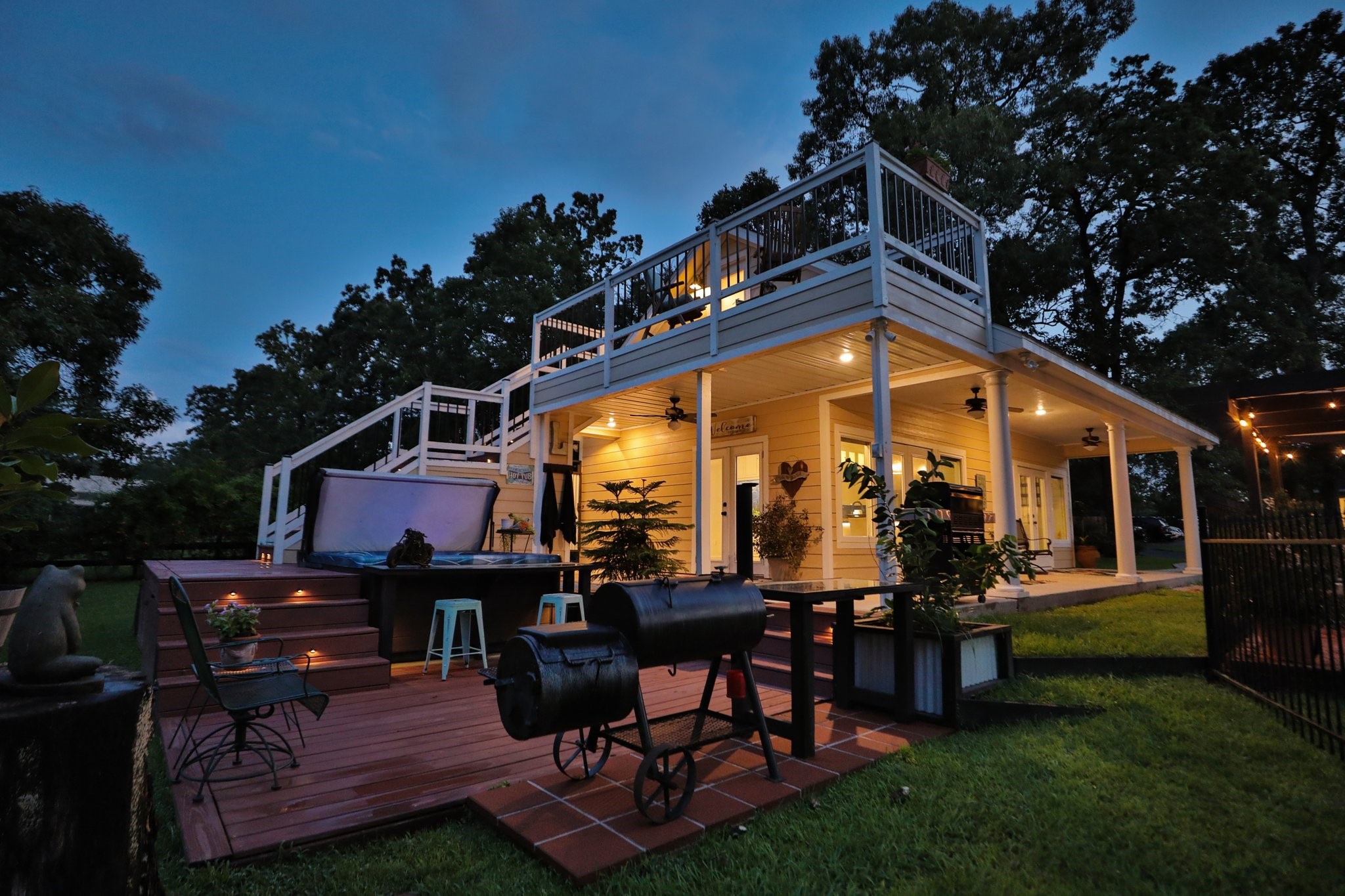 Inviting sunset view of the 1,000 guest home features a spacious two-level deck with ample outdoor seating, a hot tub, and a grill area, perfect for entertaining. The well-lit patio and surrounding greenery create a cozy ambiance for evening gatherings.