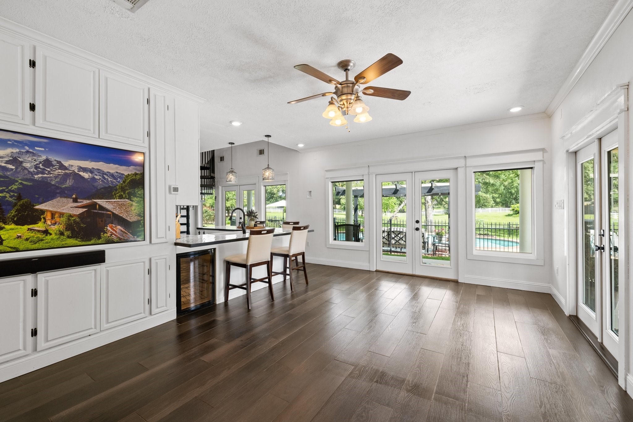 This spacious, bright living room in the guest house features elegant tile wood look floors and a ceiling fan. It opens to a scenic backyard with large windows and French doors, offering views of a pool and greenery. The open concept includes a modern kitchen with a breakfast bar and stylish lighting.