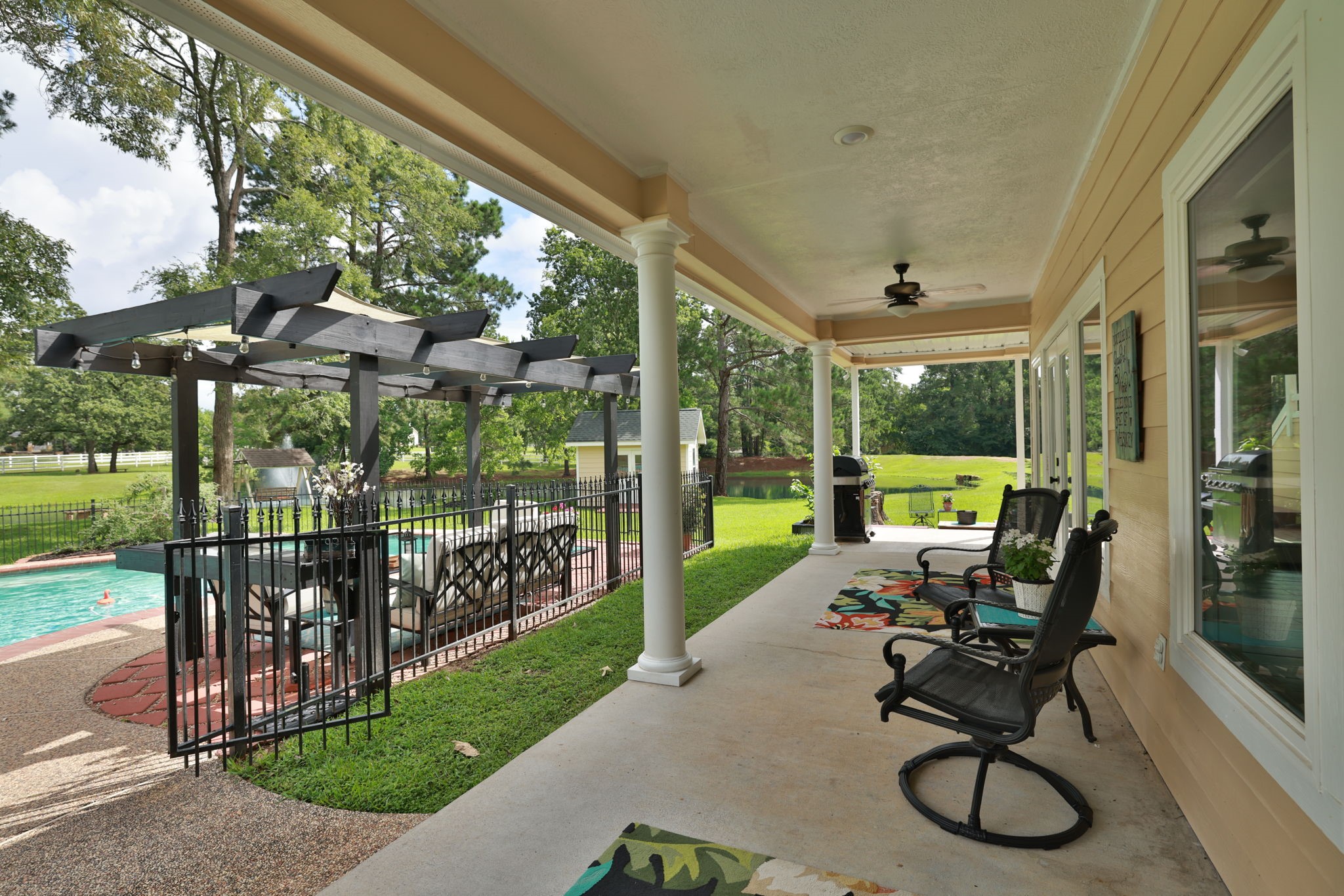 This photo showcases a spacious covered patio with ceiling fans, overlooking a fenced pool area with a pergola and seating. It offers a serene view of a lush green yard, perfect for outdoor relaxation and entertaining.