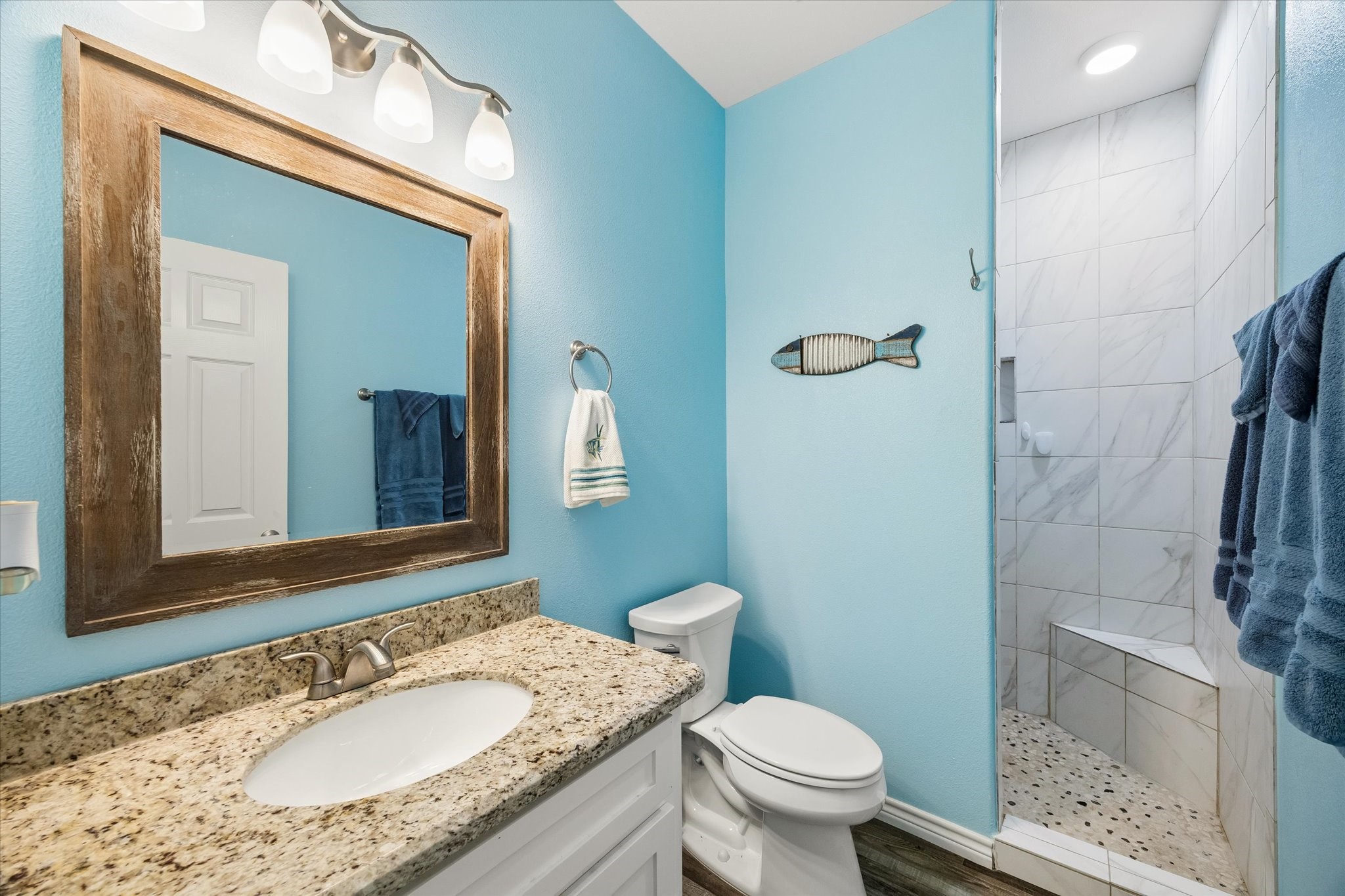 Charming bathroom with a soothing blue color scheme, featuring a granite countertop, rustic wood-framed mirror, and a stylish tiled shower with a built-in bench.