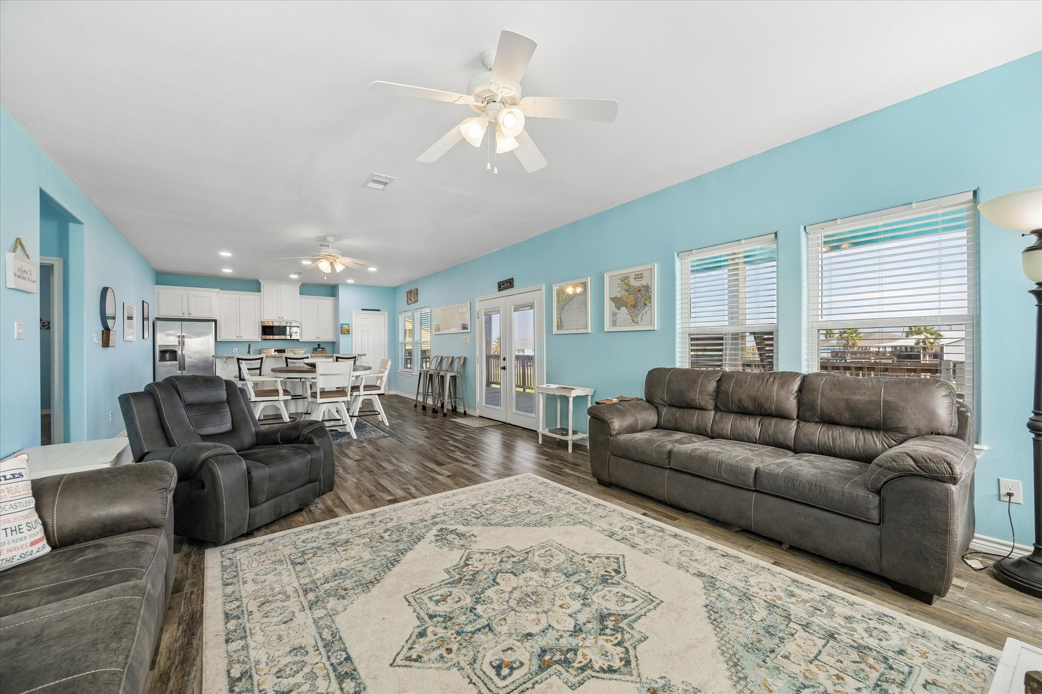 Spacious open-concept living area with vibrant blue walls, modern ceiling fans, and ample natural light. The room features seamless flow into a contemporary kitchen with sleek white cabinetry and a breakfast bar.