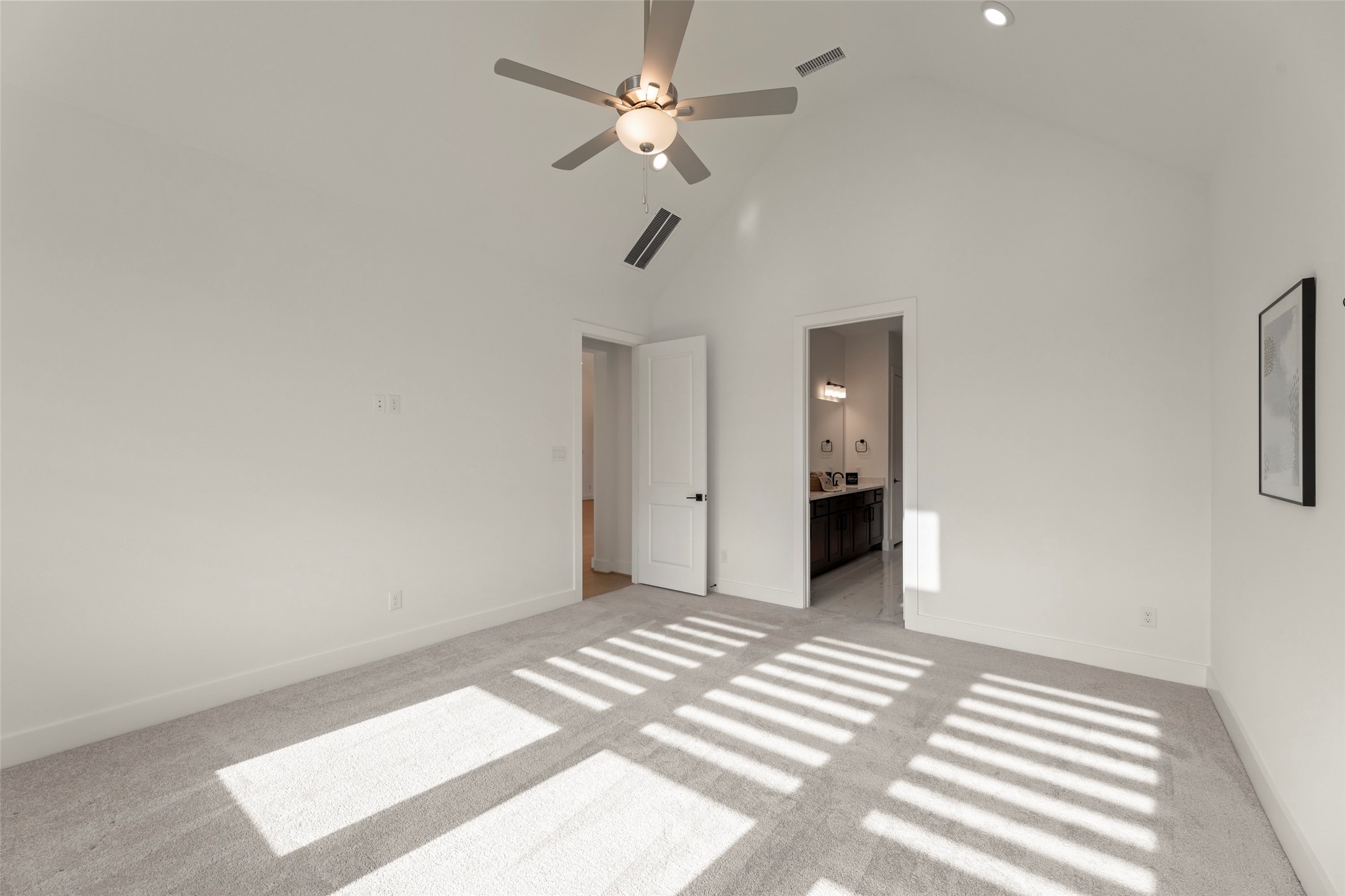 Sunlit bedroom with a vaulted ceiling, modern ceiling fan, and plush carpeting, leading to a sleek ensuite bathroom.