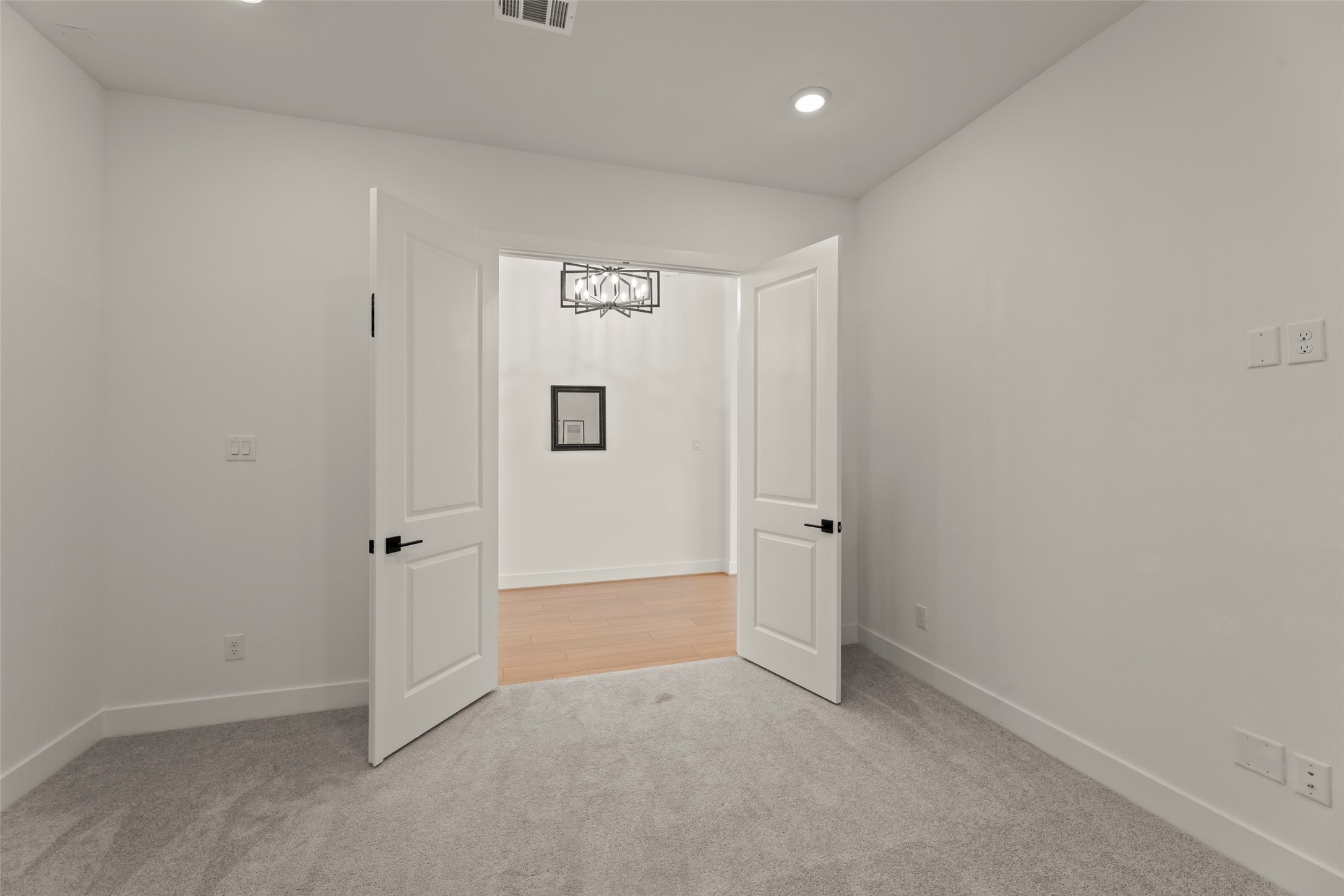 Bright room featuring plush carpeting and modern double doors leading to a hardwood-floored area, highlighted by a stylish light fixture.