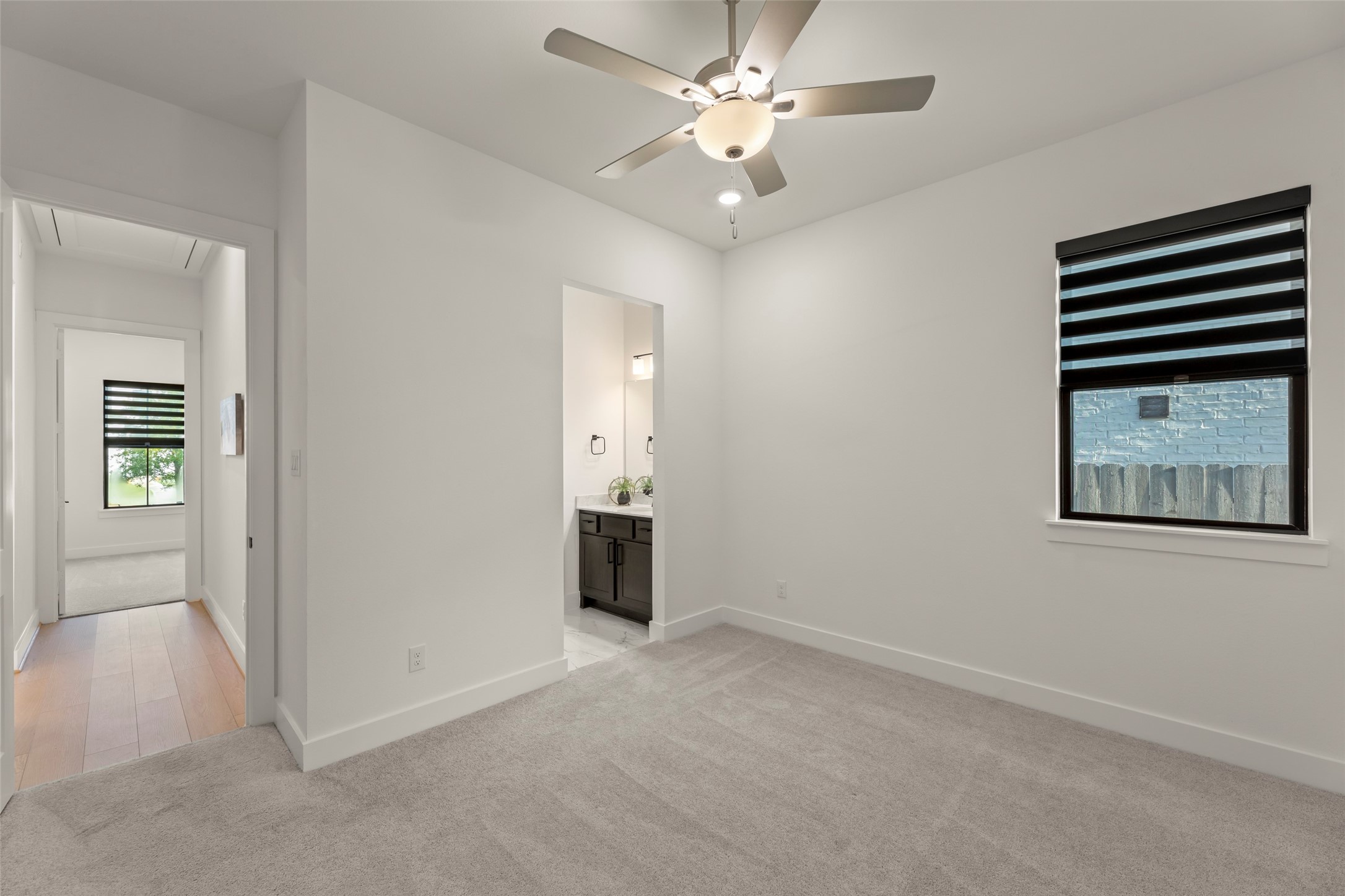 Welcoming bedroom featuring plush carpeting, a ceiling fan, and a window with modern blinds. The room connects to a hallway and an adjoining bathroom with sleek fixtures.