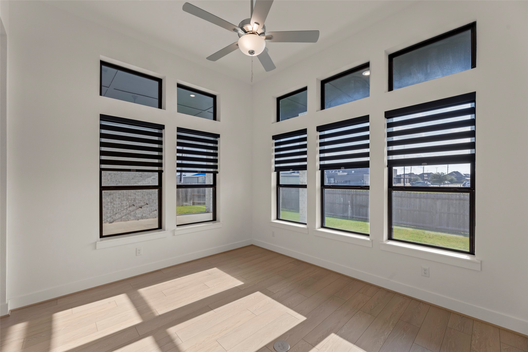 Airy room featuring large windows with modern blinds, light wood flooring, and a ceiling fan, perfect for relaxation or a home office setup.