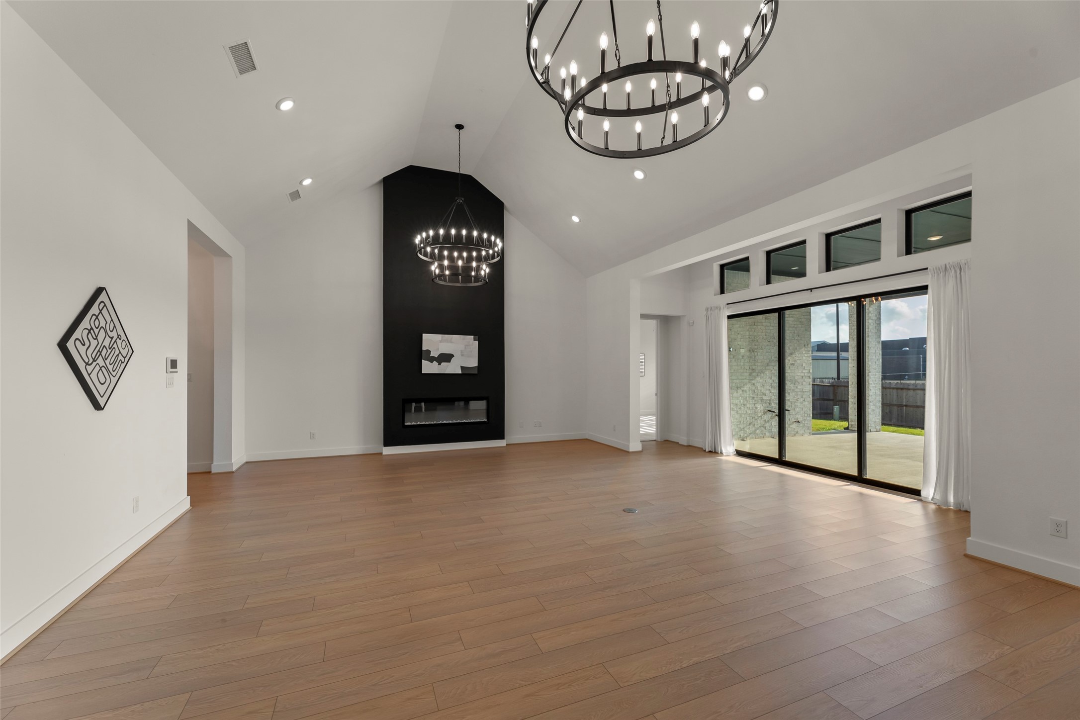Vaulted ceilings, elegant chandeliers, and a modern black accent wall featuring a fireplace. Expansive windows and sliding glass doors offer ample natural light and access to a patio.