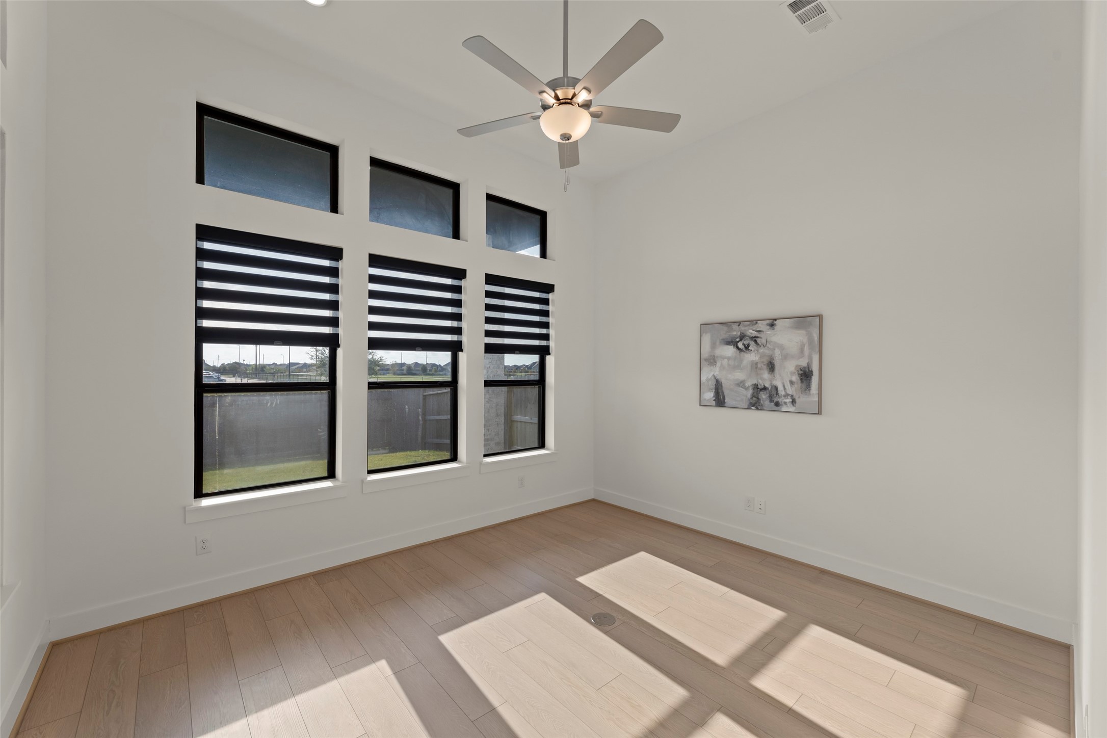 Inviting room featuring large windows with modern blinds, sleek ceiling fan, and light wood flooring, perfect for a tranquil retreat.