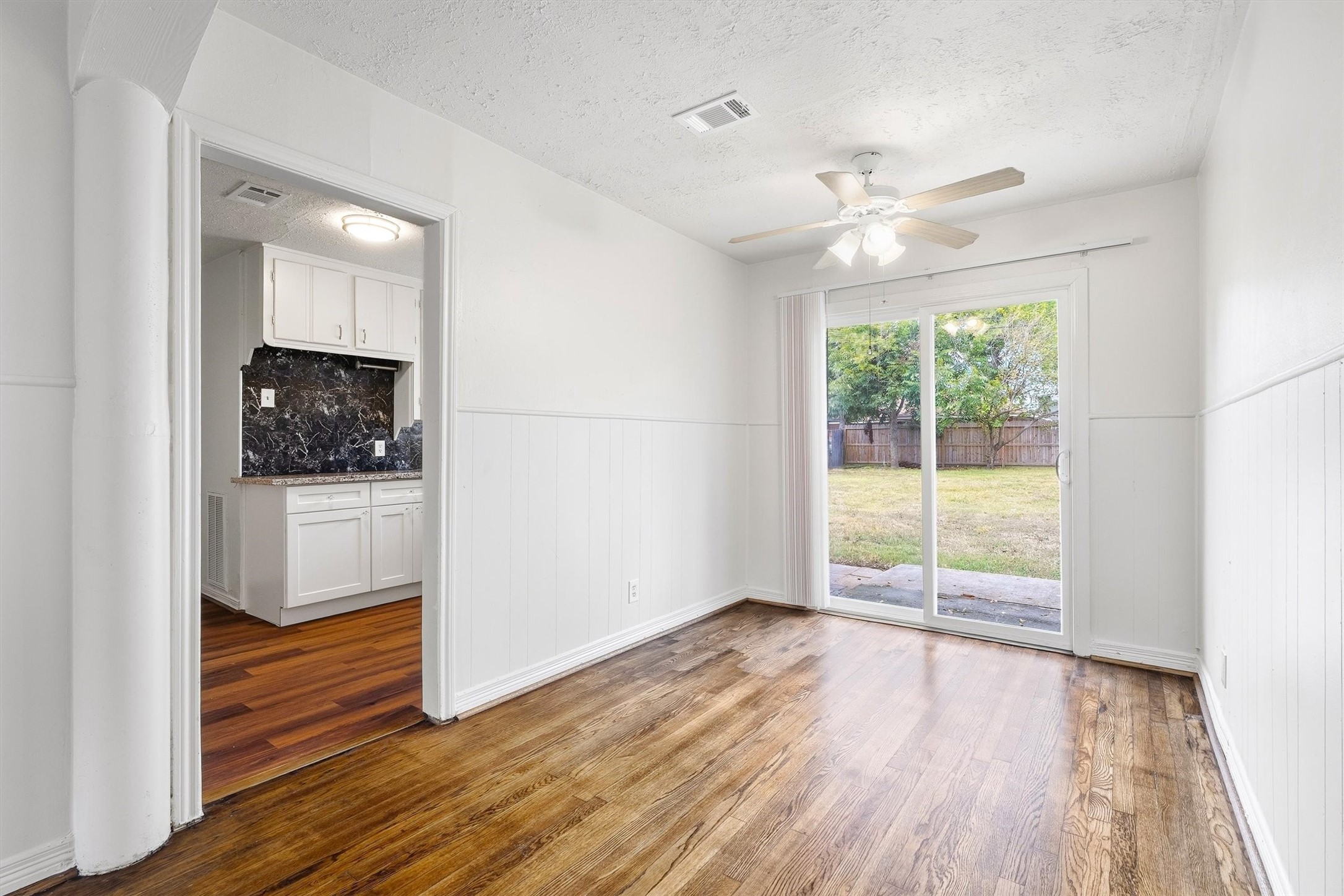 Bright dining area with hardwood floors, sliding glass doors leading to a spacious backyard, and an adjacent kitchen featuring marble-patterned backsplash.