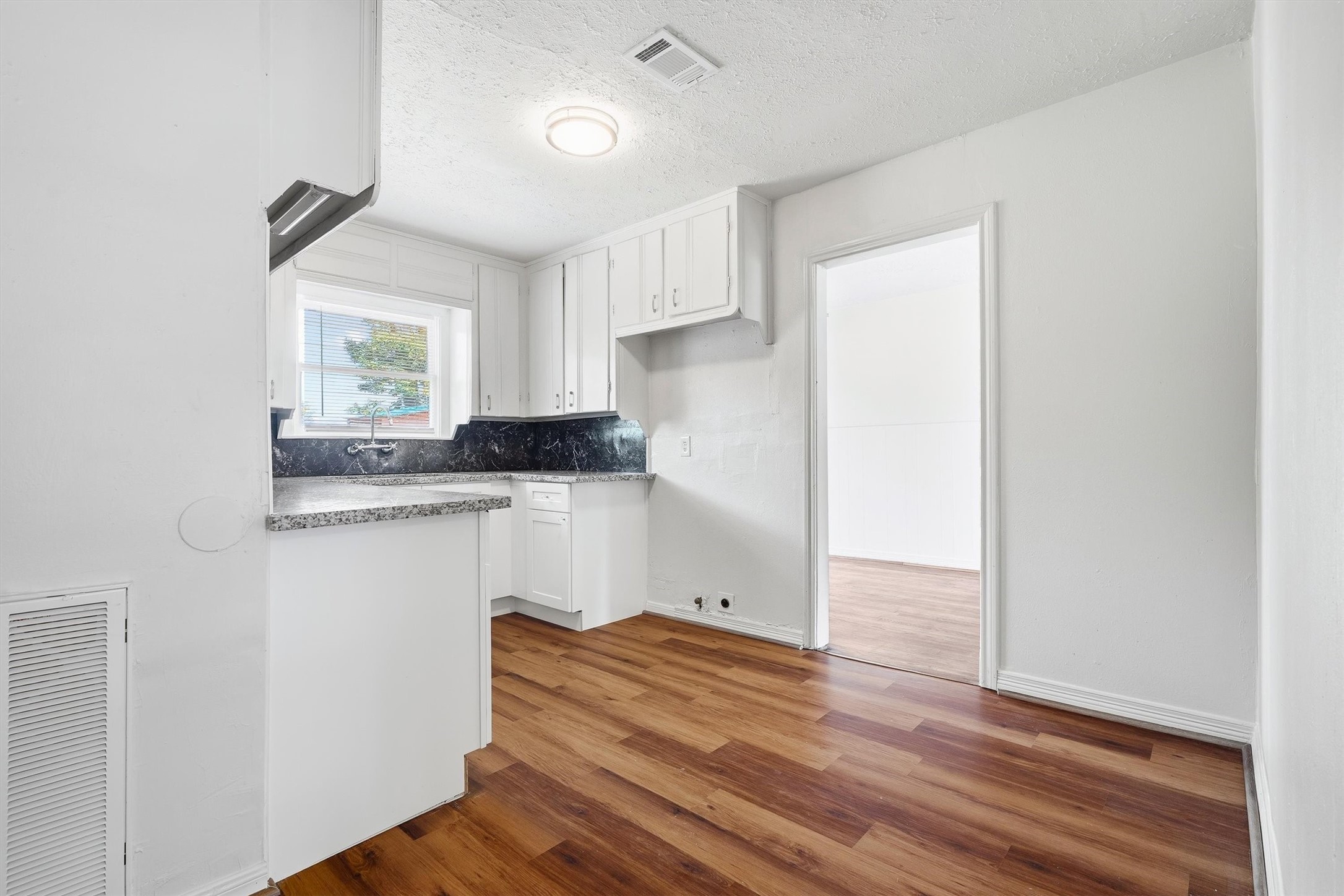 Charming kitchen with rich hardwood floors, sleek granite countertops, and ample white cabinetry, leading into a bright adjoining room.