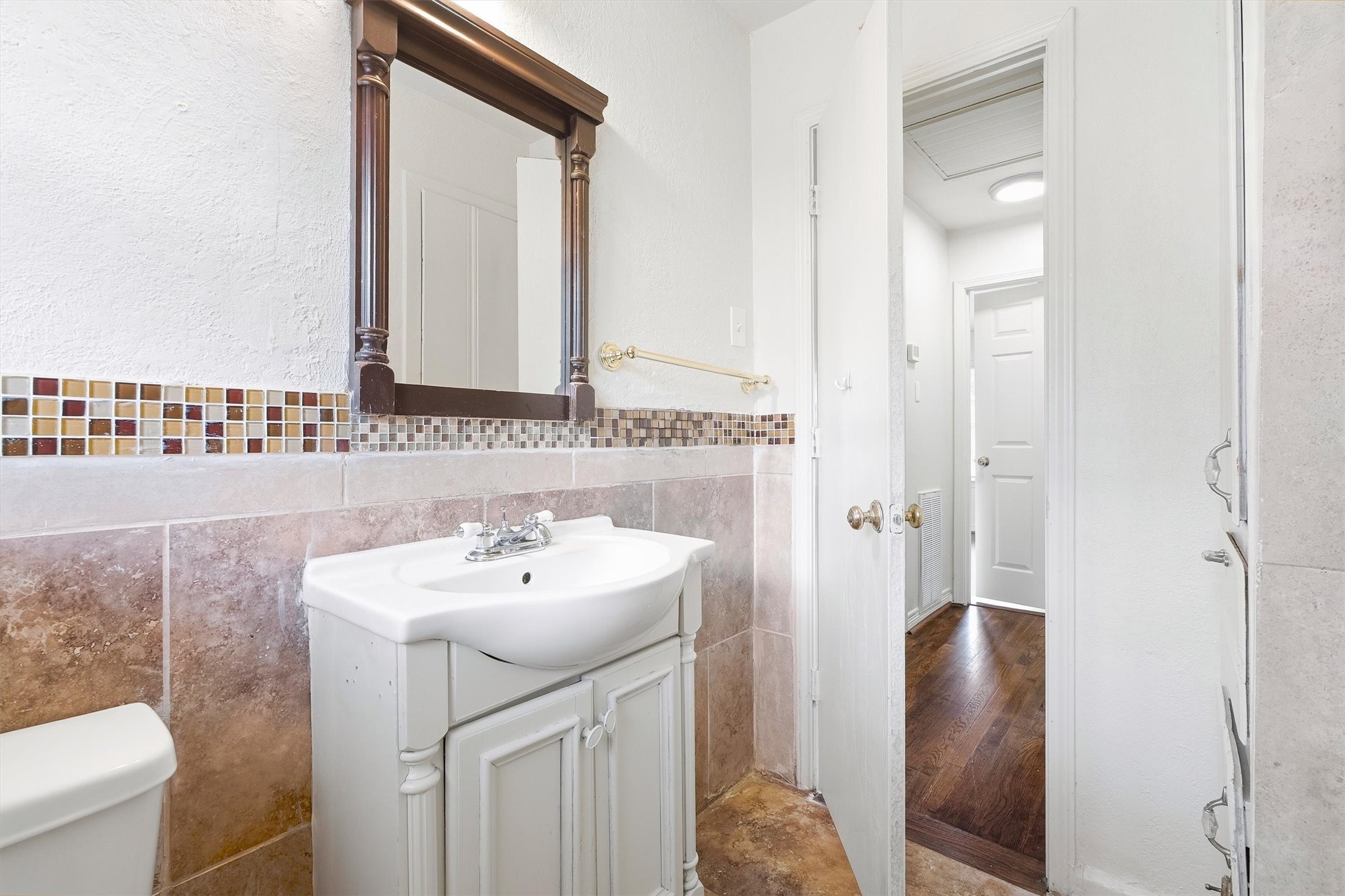 Nice bathroom featuring a classic vanity with a decorative mirror, tile backsplash, and warm-toned flooring leading to a bright hallway.